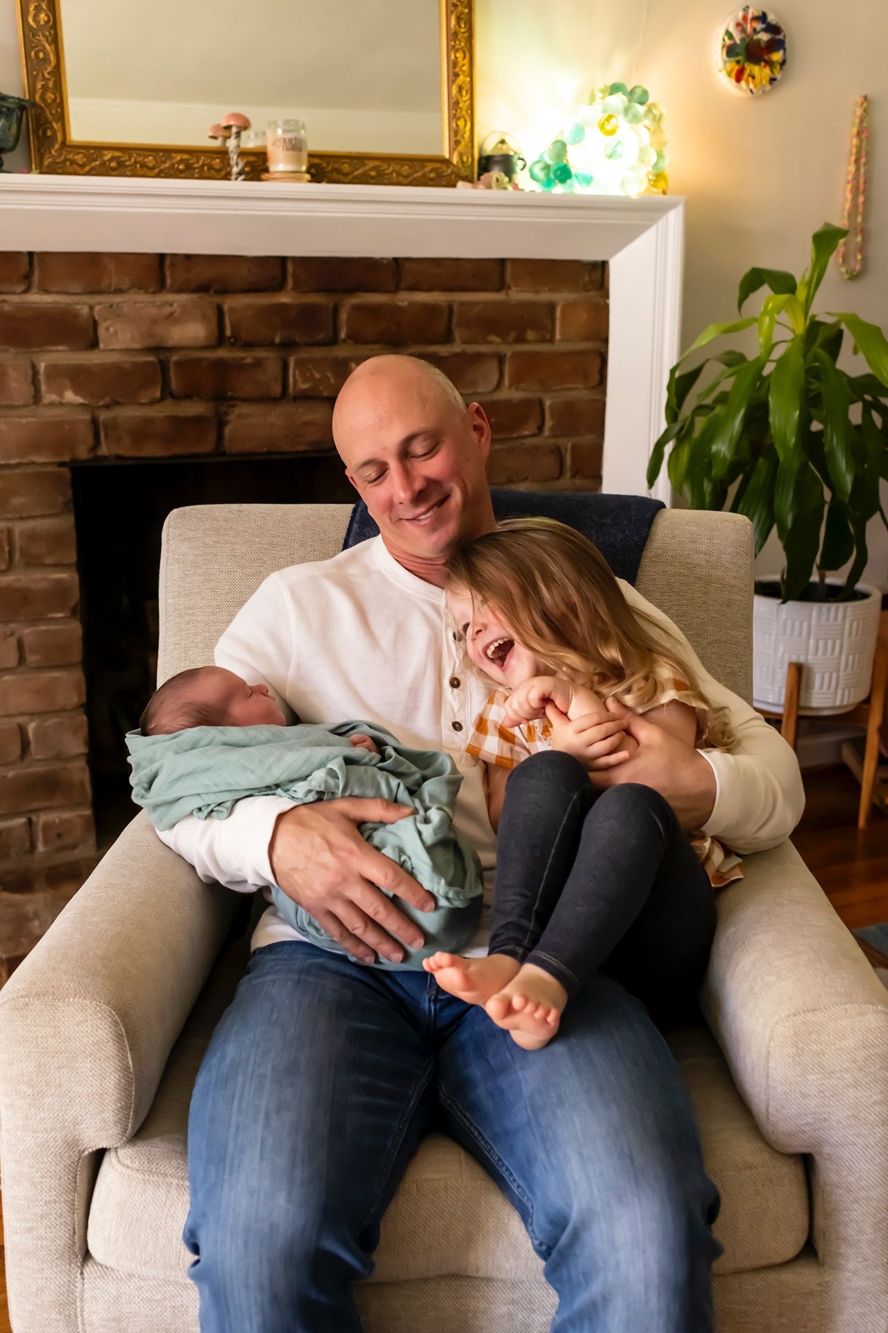 Parent and child cuddling on armchair with newborn baby in front of brick fireplace.