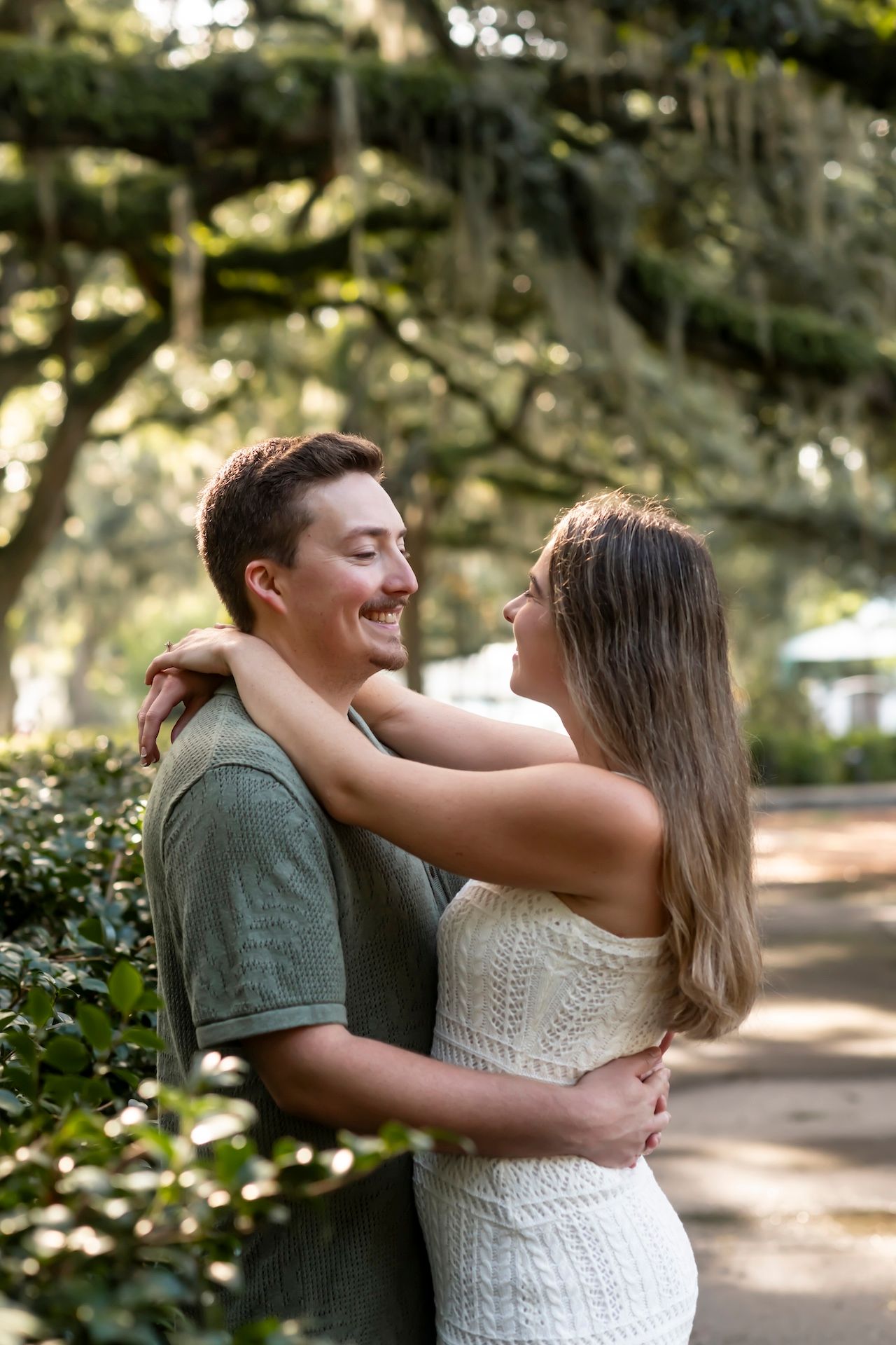 A romantic couple embraces on a sunlit path beneath Spanish moss draped trees in a romantic outdoor setting.