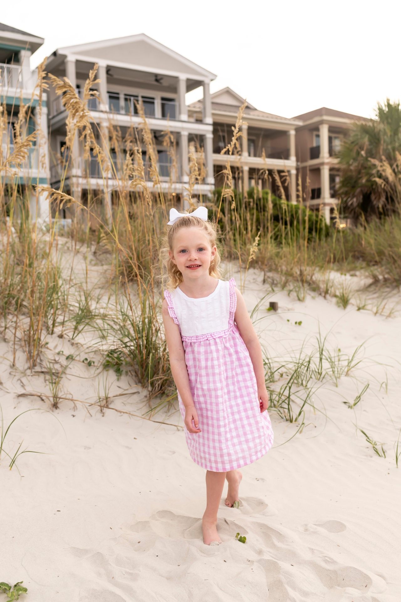 A child in a pink gingham dress plays happily on a sandy beach near beach houses and sea oats.