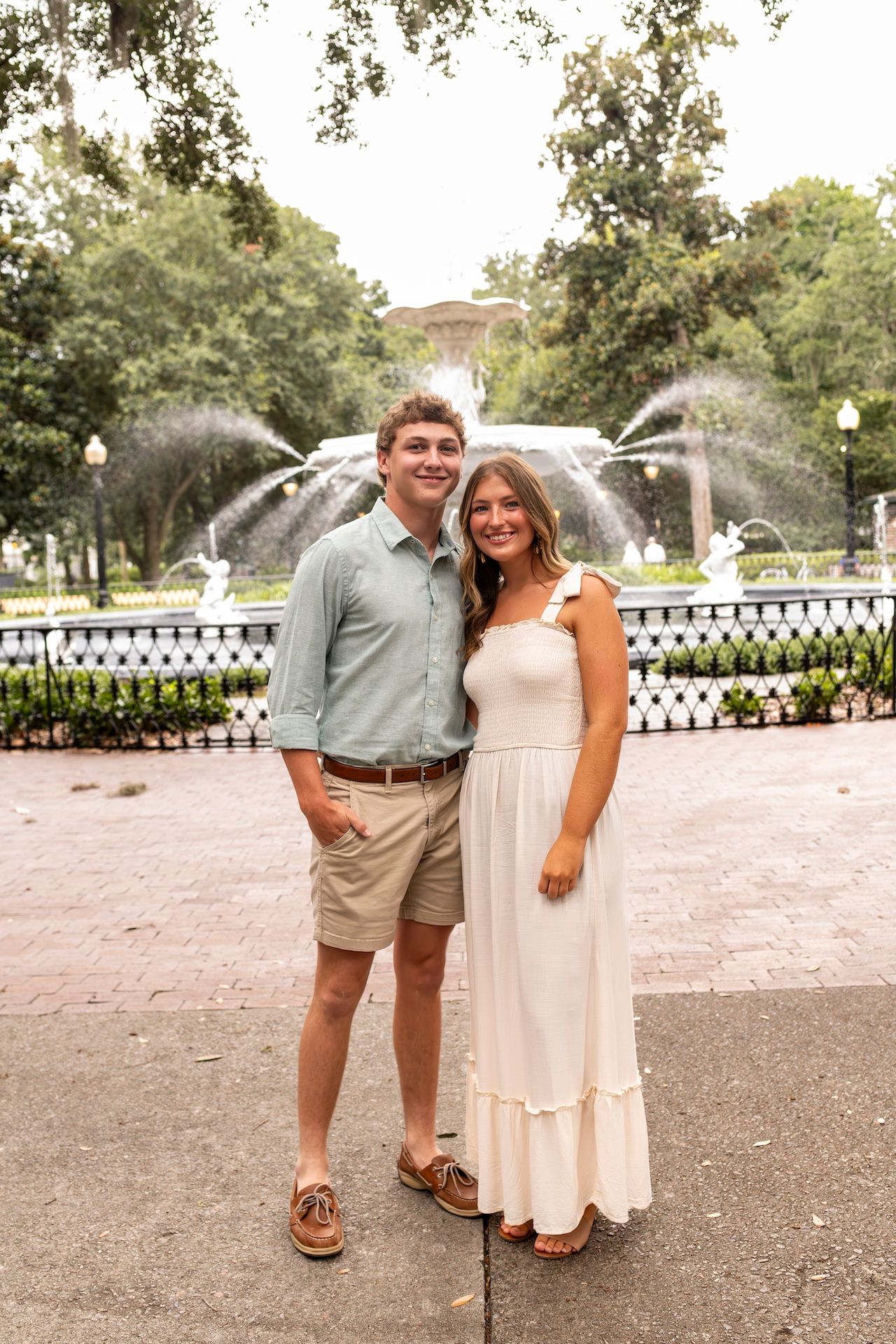 Elegant couple poses by a decorative fountain in a park with trees and brick pavers.