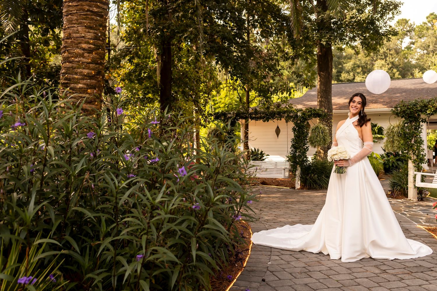 A bride in a flowing white wedding dress stands on a garden path surrounded by lush greenery and palm trees at sunset.