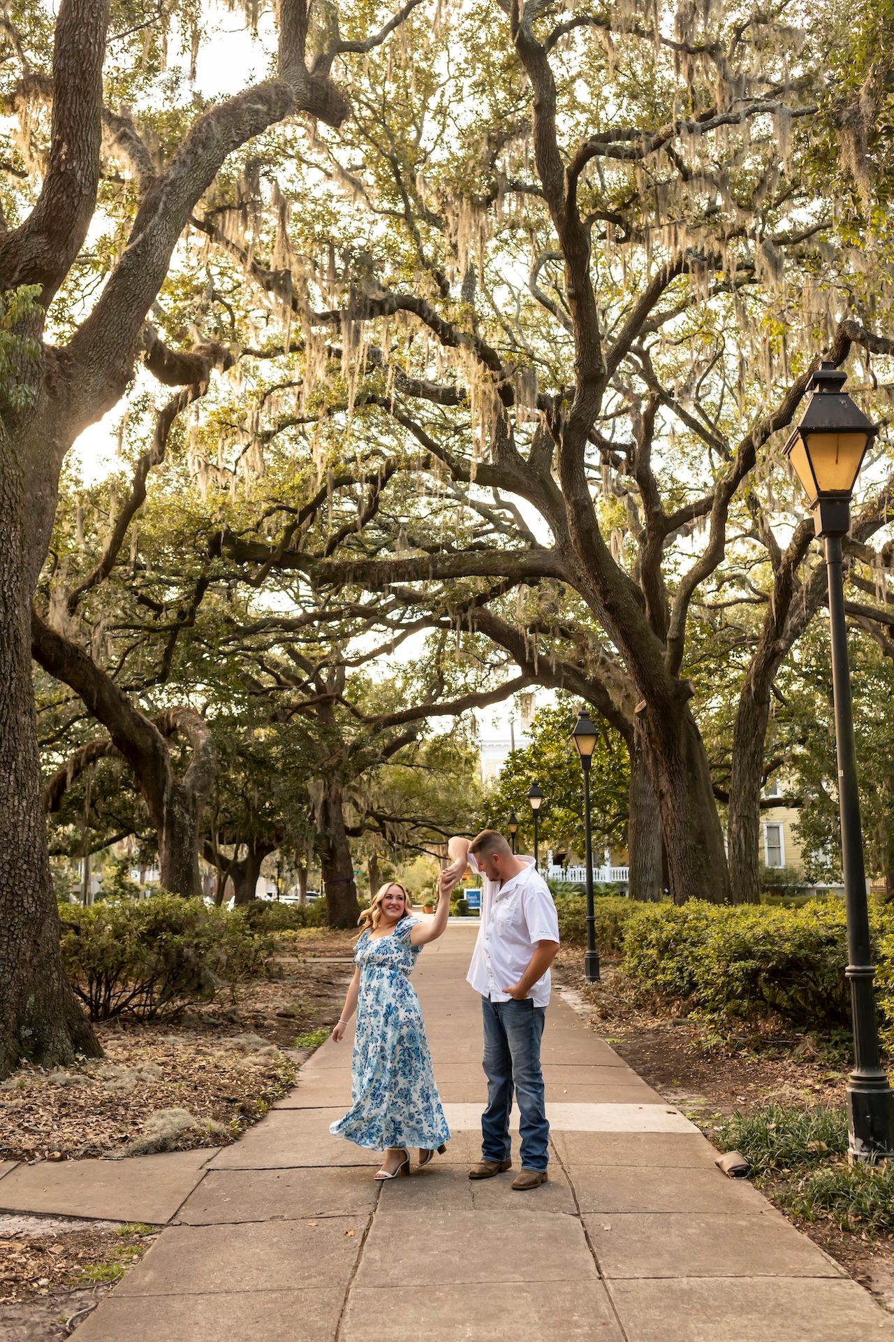 A couple stands on a scenic pathway under large oak trees with hanging moss.