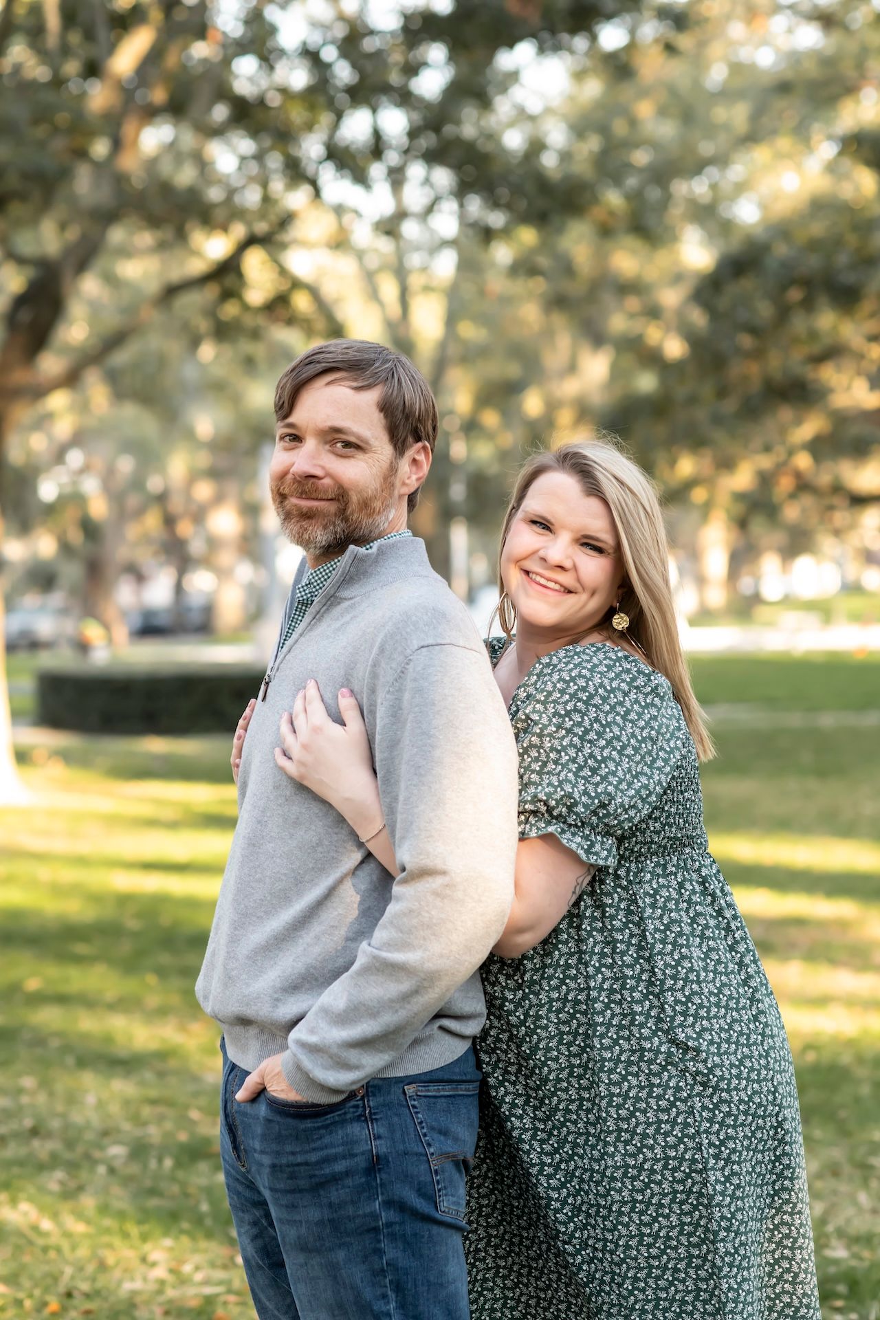 A couple embraces outdoors in a sunlit park, with the woman in a patterned dress hugging the man in a gray sweater from behind.
