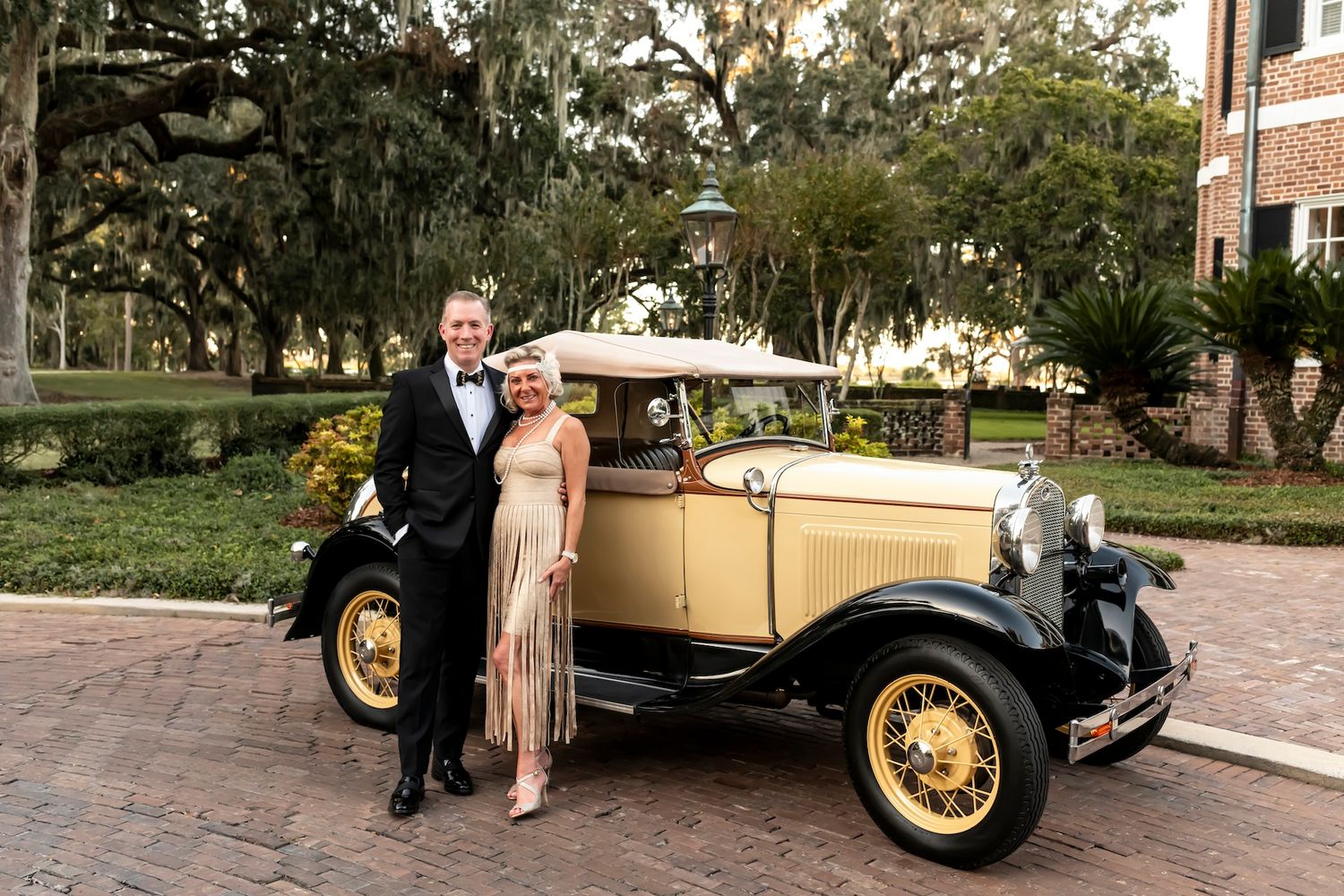 A vintage cream-colored Model A Ford parked on brick pavement beside Spanish moss draped trees.