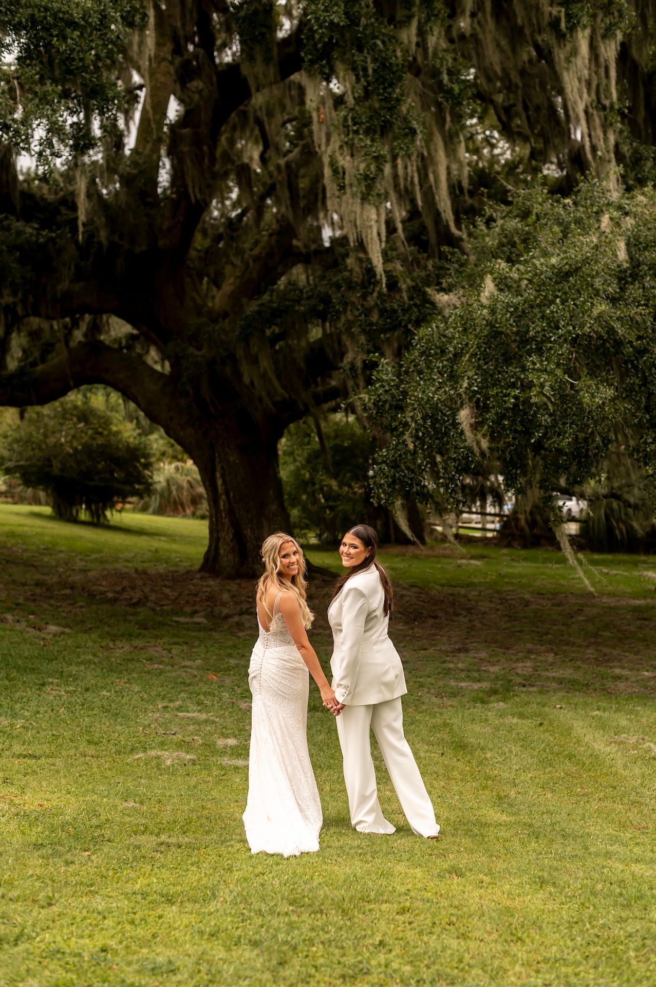 Two people in wedding attire hold hands under a majestic oak tree draped with Spanish moss in a Southern garden setting.