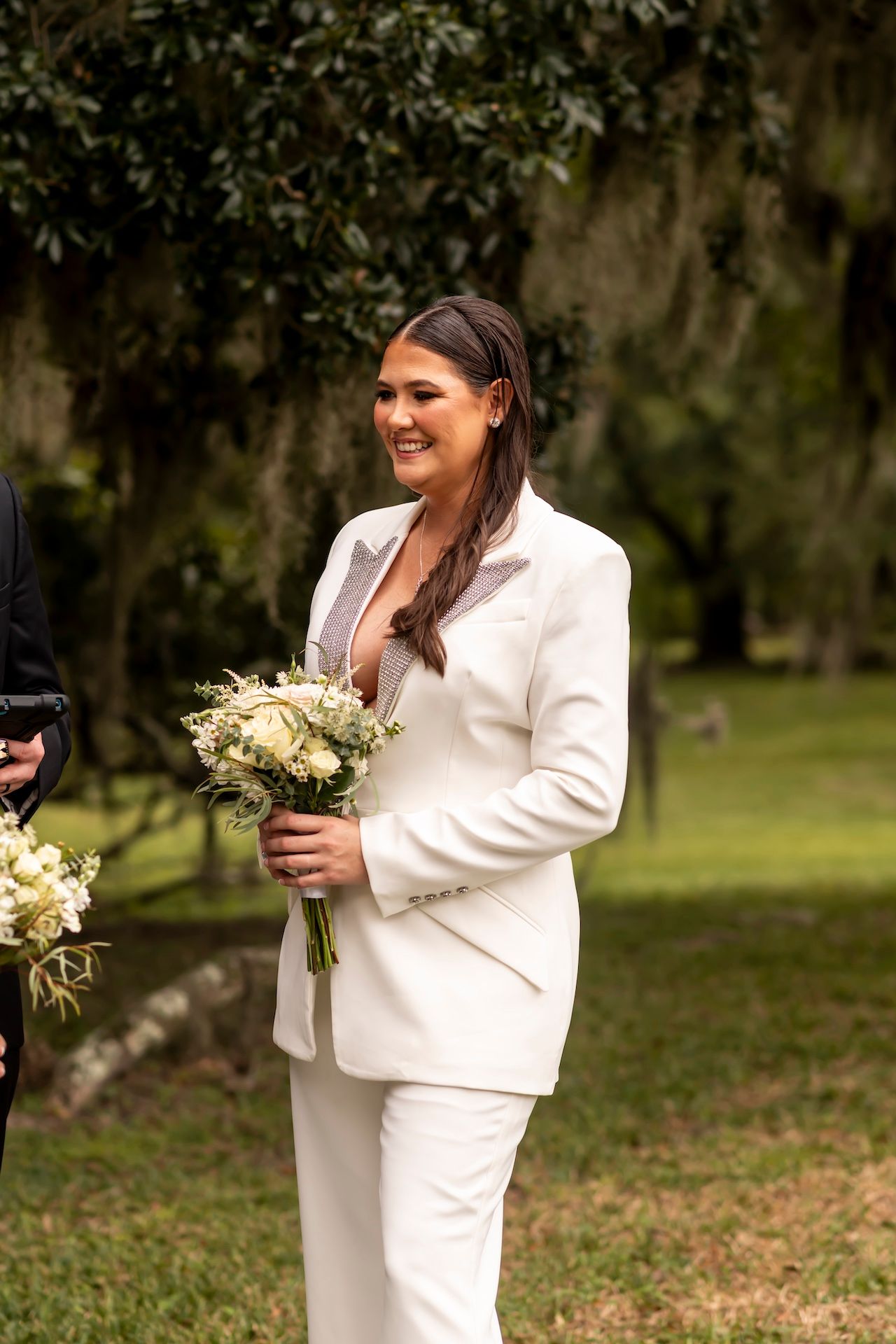 Someone in an elegant white suit holds a delicate bouquet of white flowers during an outdoor ceremony.