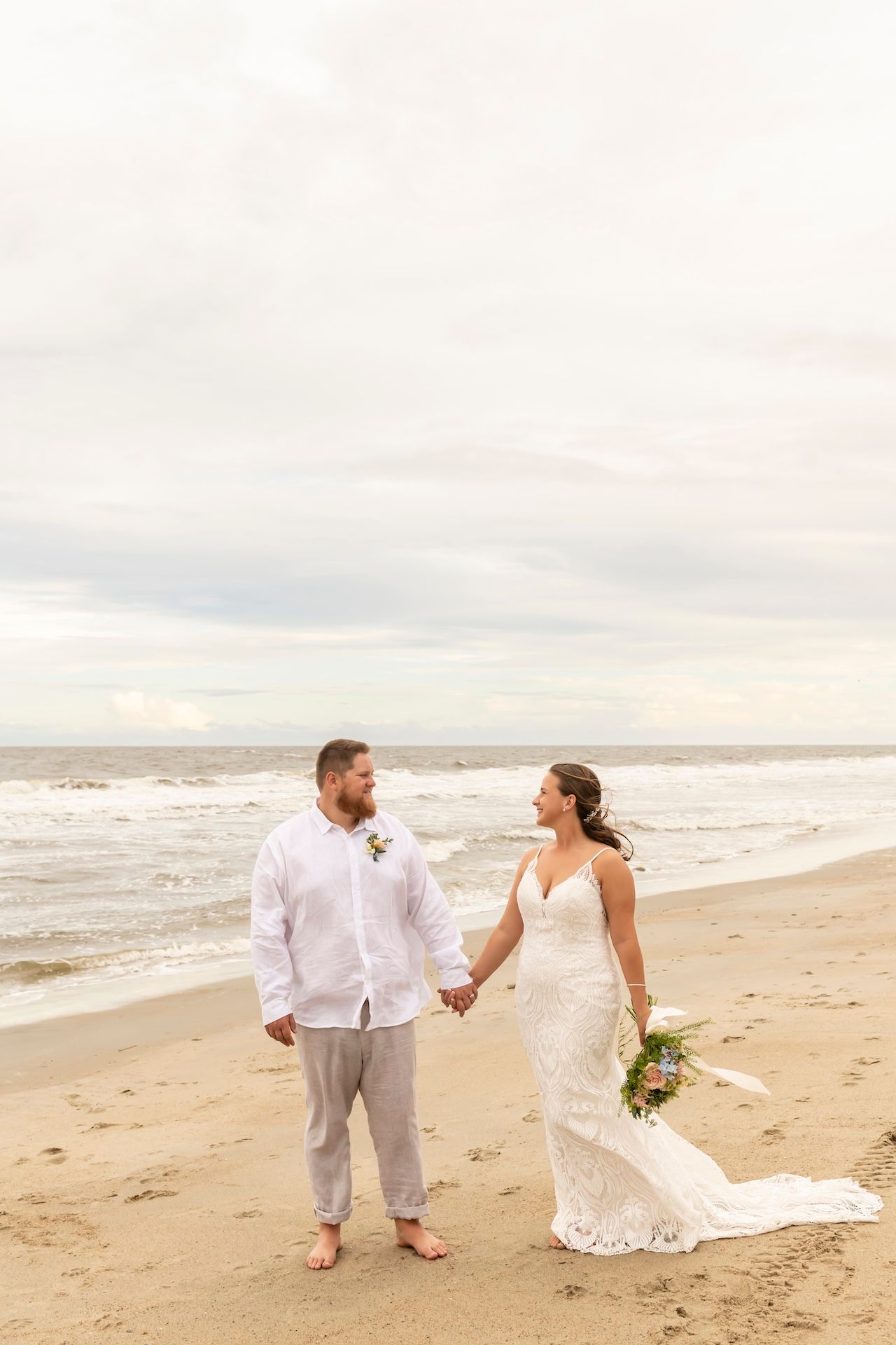 Couple enjoying romantic beach wedding photoshoot on sandy shore with crashing waves and overcast sky.