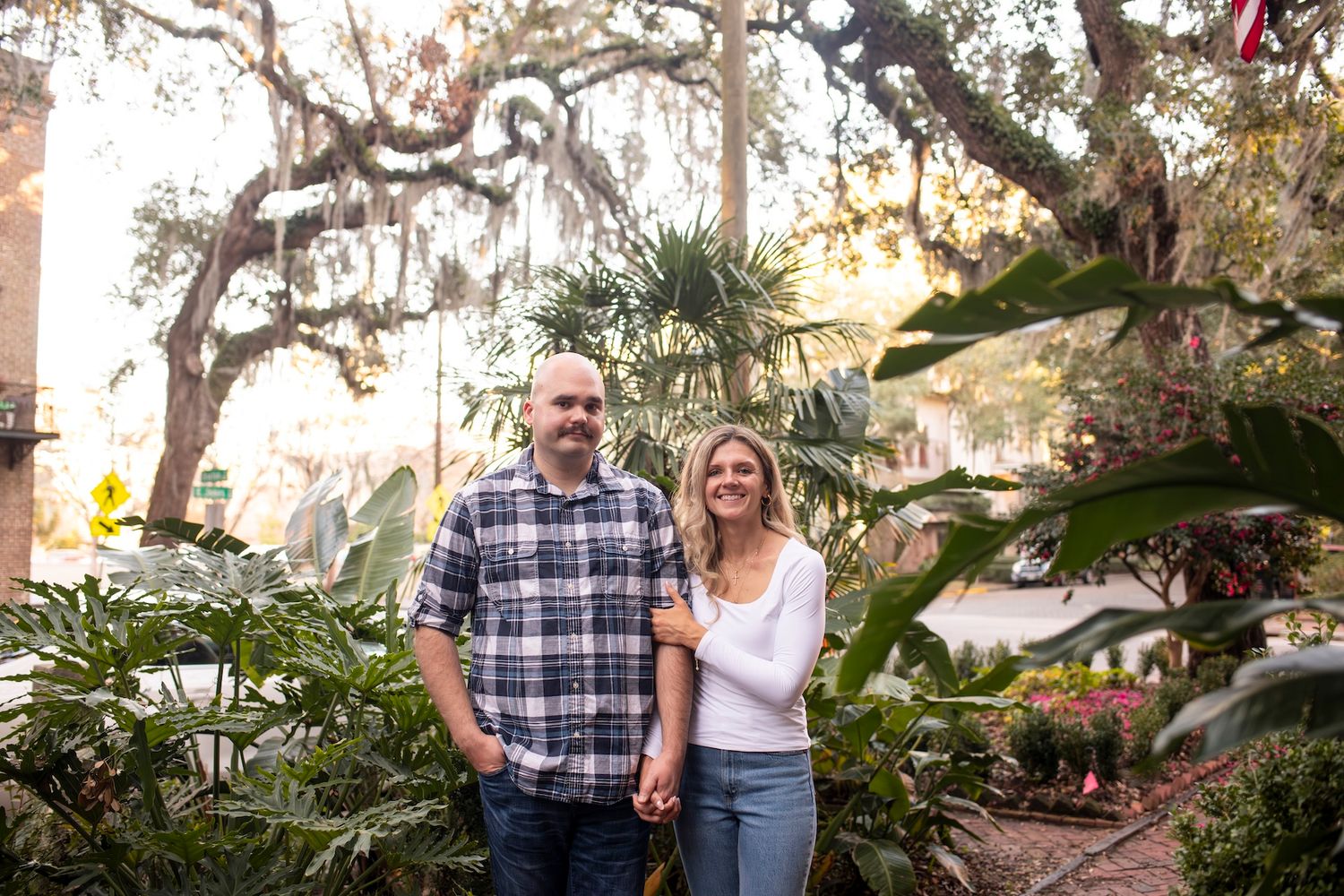 A couple poses together outdoors on a sunny day with Spanish moss hanging from trees in the background creating a romantic scene.