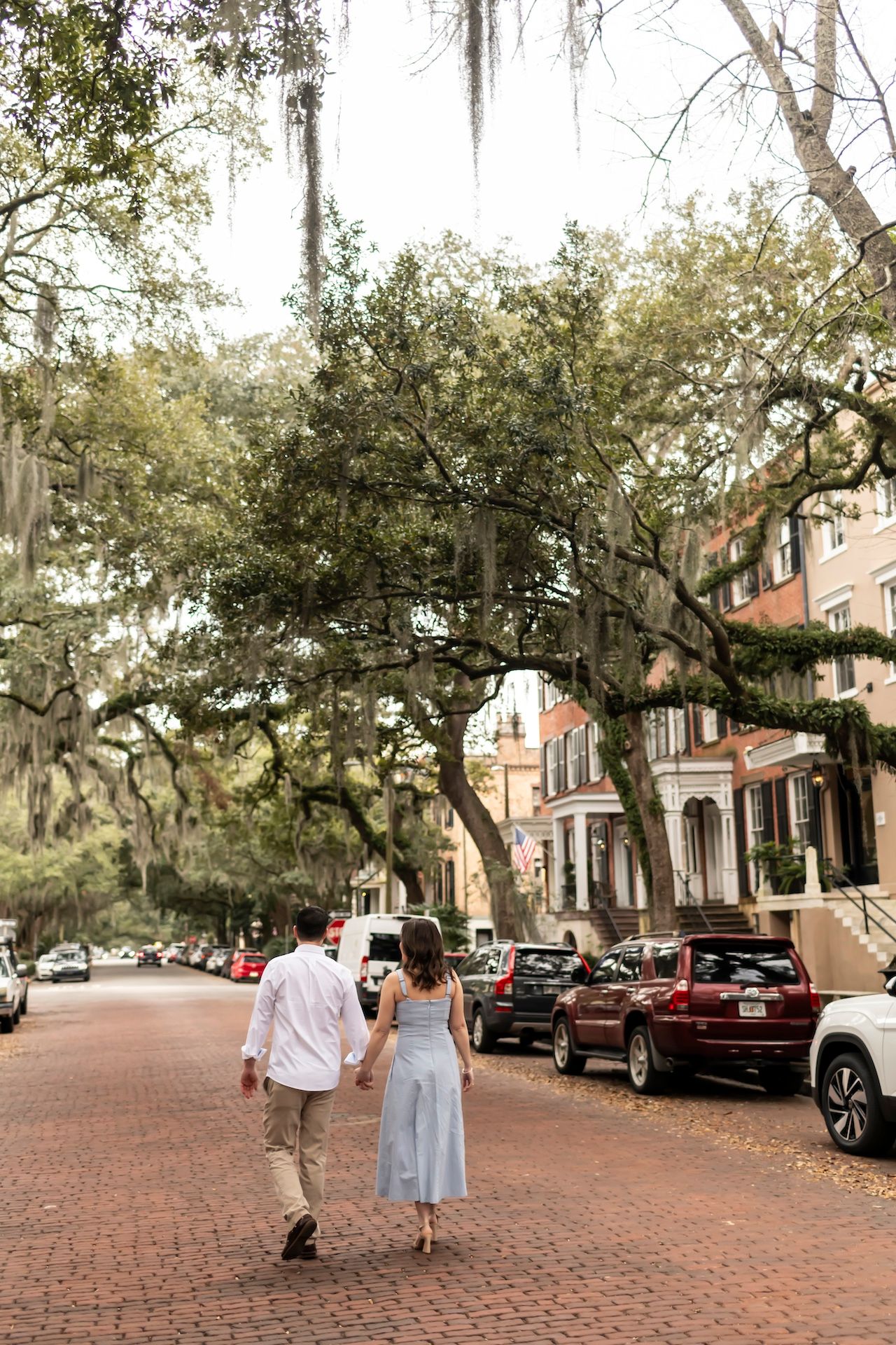 Couple walking down brick sidewalk beneath Spanish moss-draped oak trees in historic Savannah Georgia.