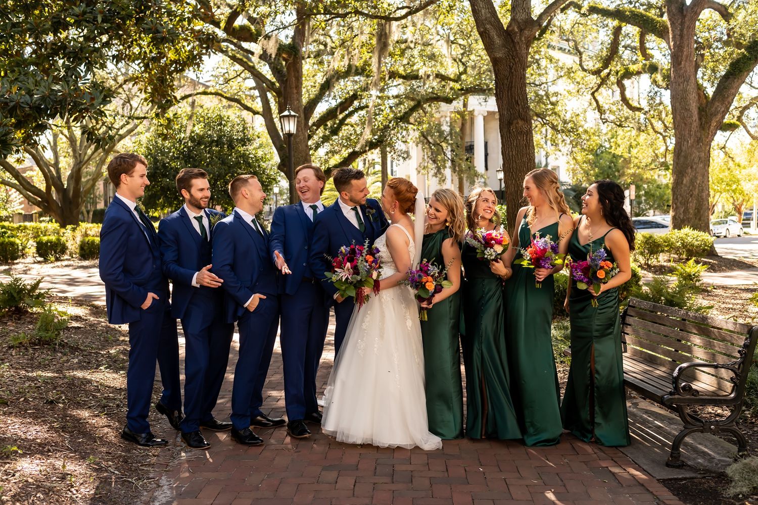 A wedding party poses together under sunlit oak trees wearing navy suits and emerald green dresses.