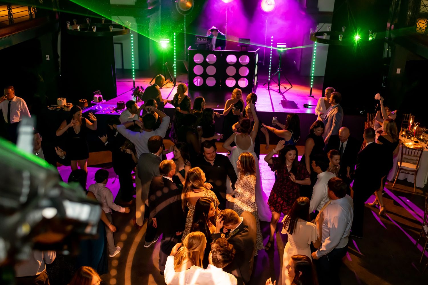 A crowded nightclub dance floor lit with vibrant purple and green lights while people dance under spotlights.