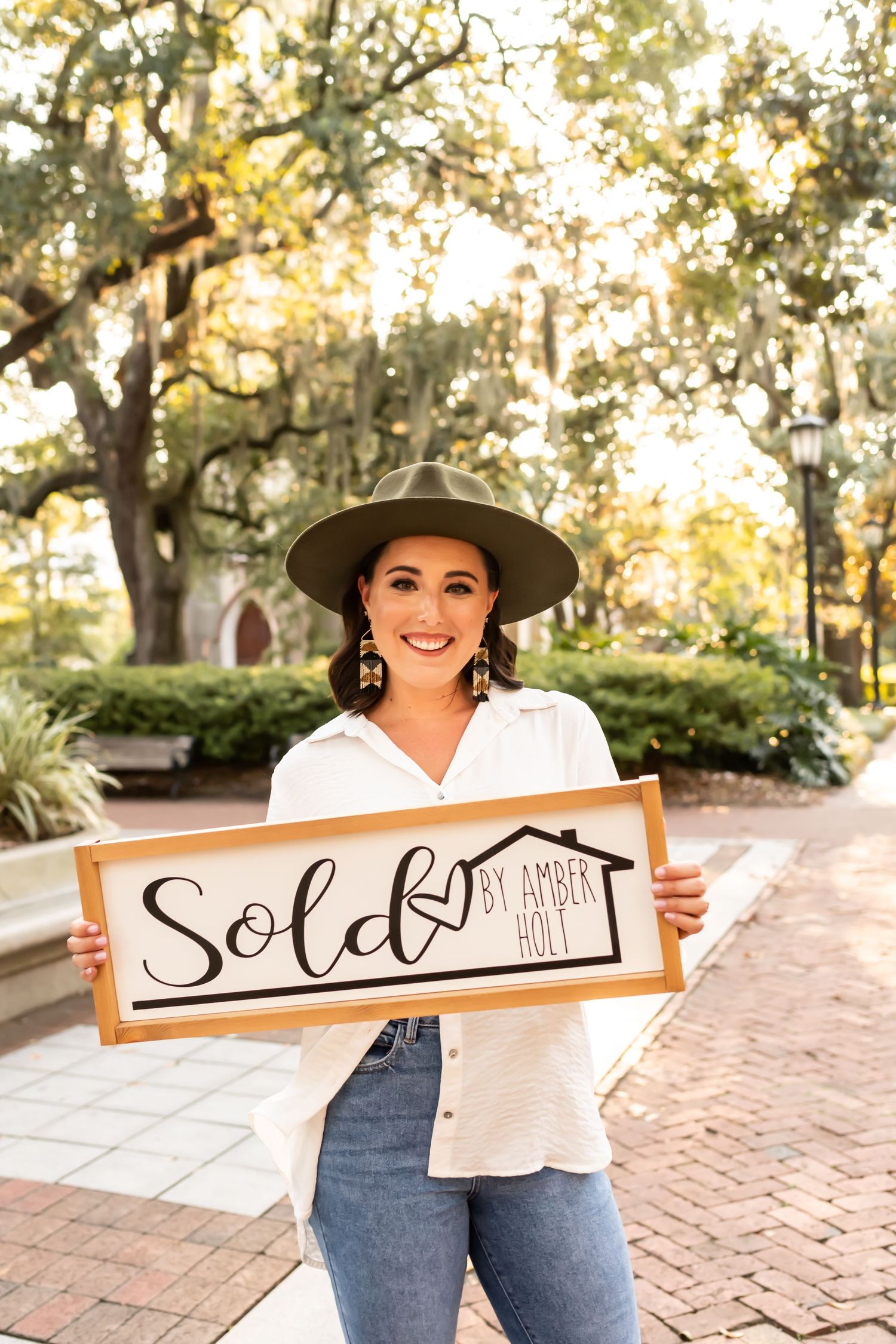 Real estate agent in jeans and white shirt holding wooden 'Sold' sign on brick pathway with trees in background.