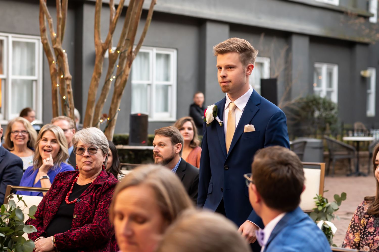 A person in a navy suit stands among seated guests at an outdoor wedding ceremony.