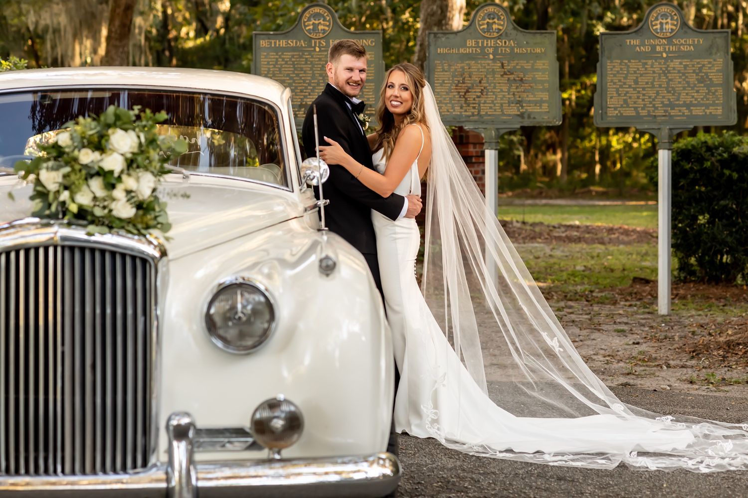 Newlyweds pose elegantly beside a classic white Rolls Royce decorated with floral arrangements at their outdoor wedding.