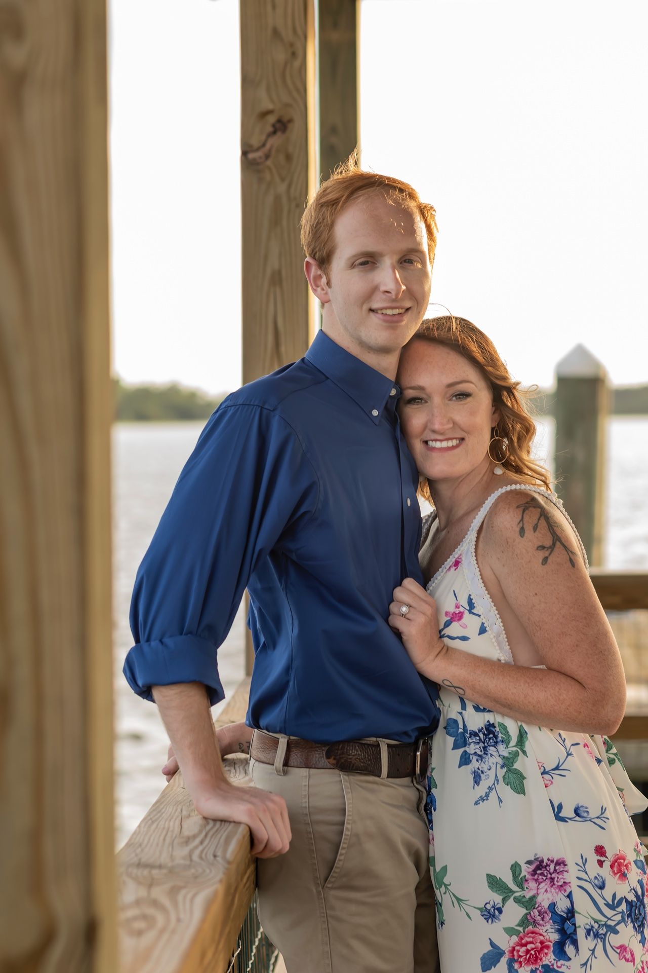 A couple poses together on a waterfront deck at sunset, with her wearing a floral dress and him in a navy blue shirt.