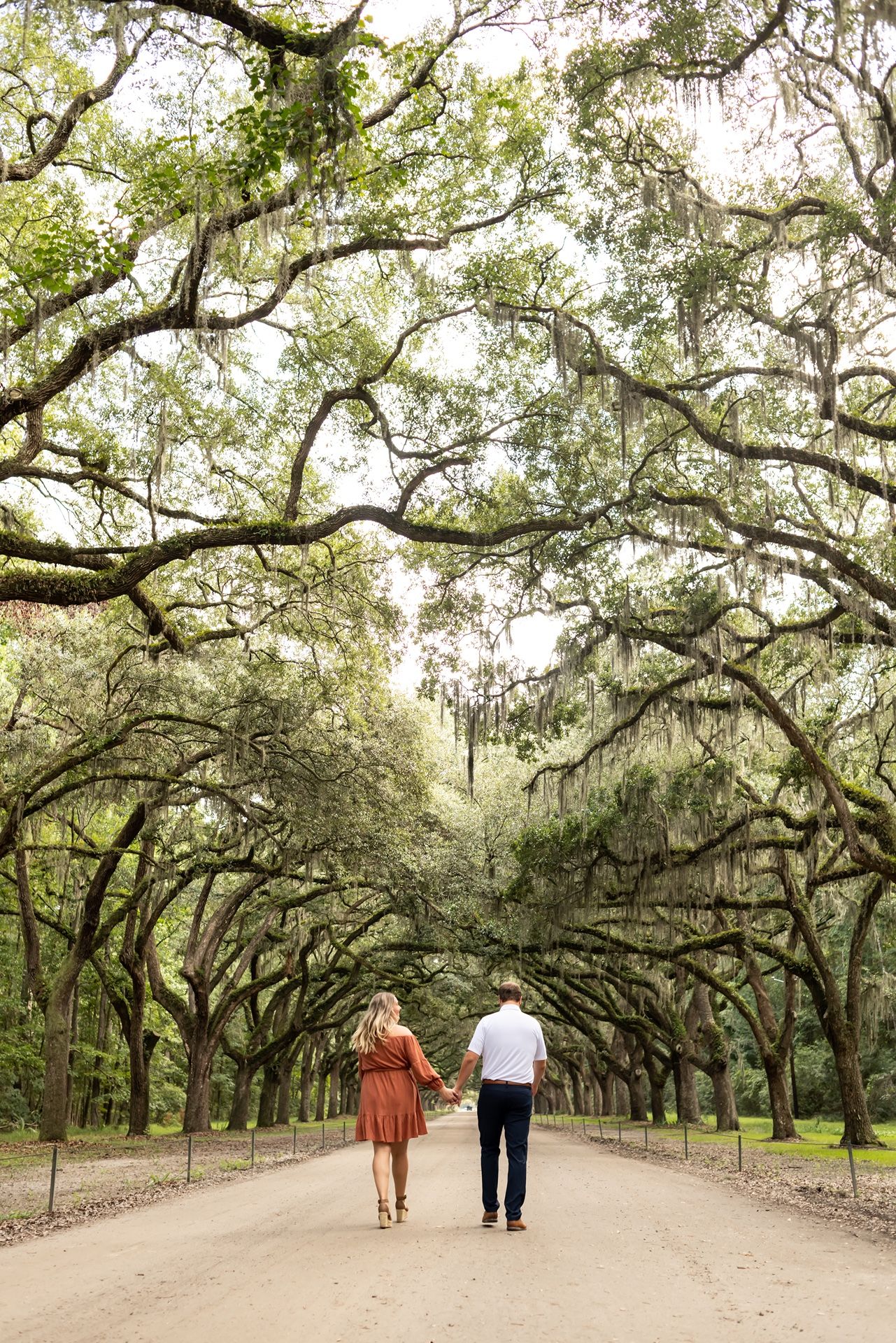 Two people walking together on a tree-lined path with Spanish moss hanging from ancient oak branches overhead.