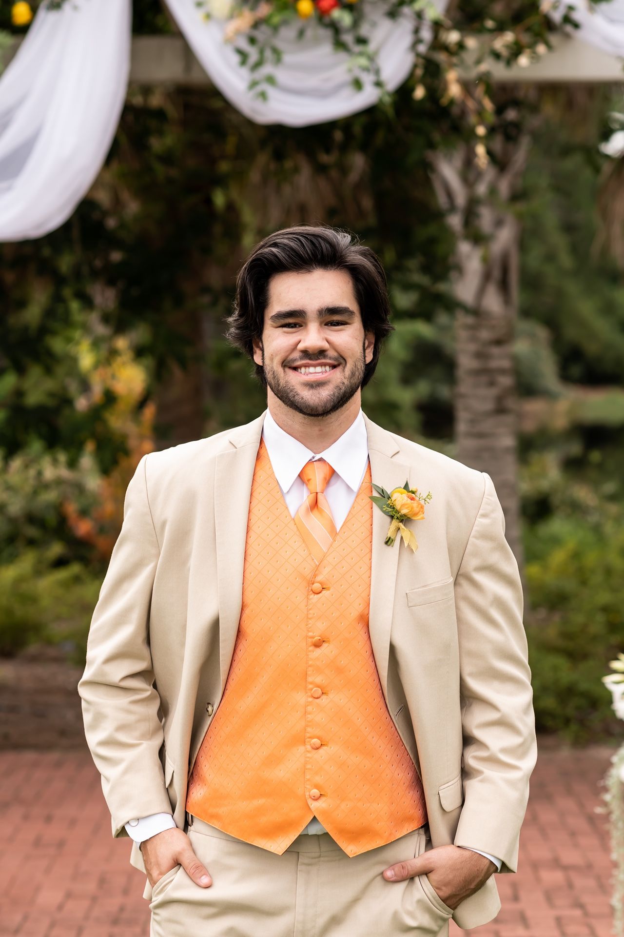 A groom wears a cream-colored suit with bright orange vest and tie at an outdoor wedding ceremony.