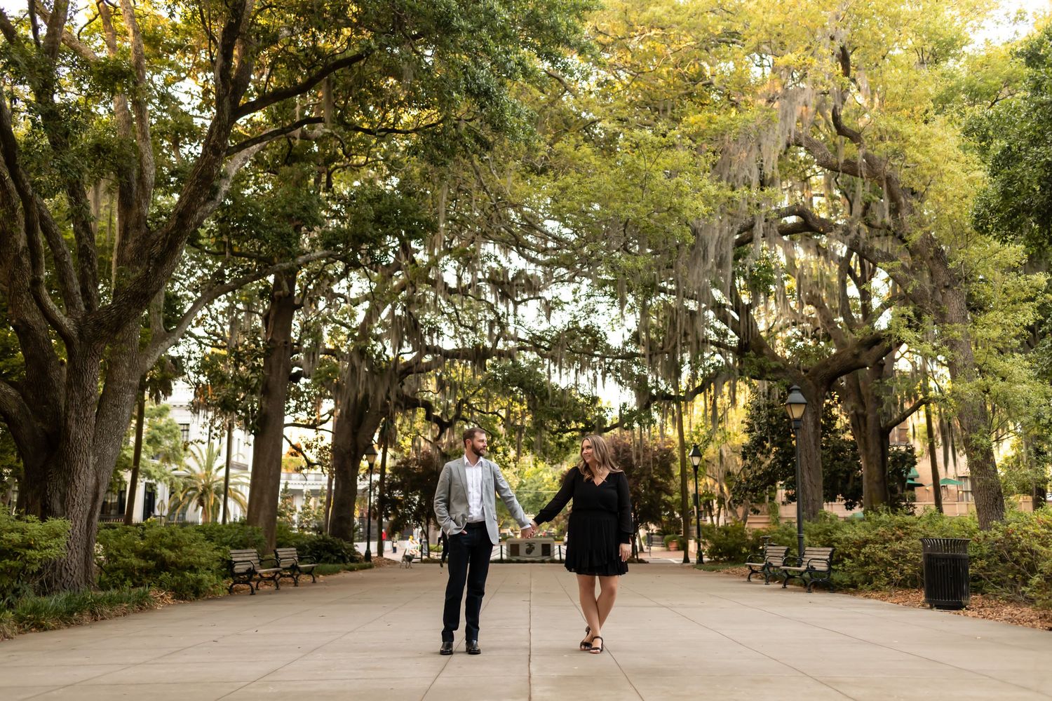 Two people stroll down a scenic path lined with Spanish moss-draped oak trees in a tranquil park setting.