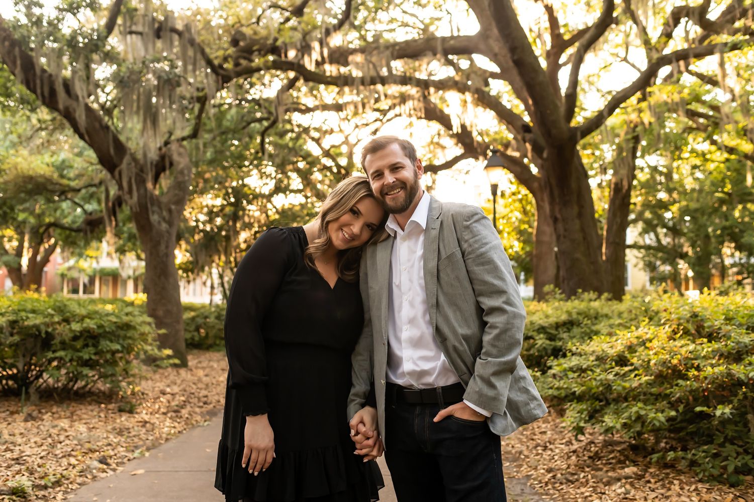 A romantic couple stands together on a path beneath Spanish moss-draped oak trees in a Southern park at sunset.