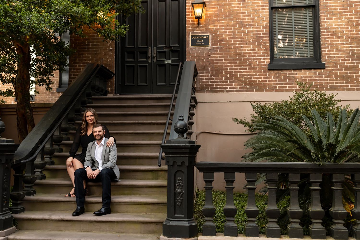 Two people sit together on steps of historic brownstone building with ornate black railing and brick facade in New York City.