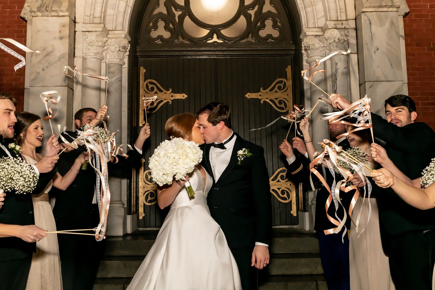 Newlyweds share a romantic kiss on church steps as wedding guests celebrate with sparklers at night.