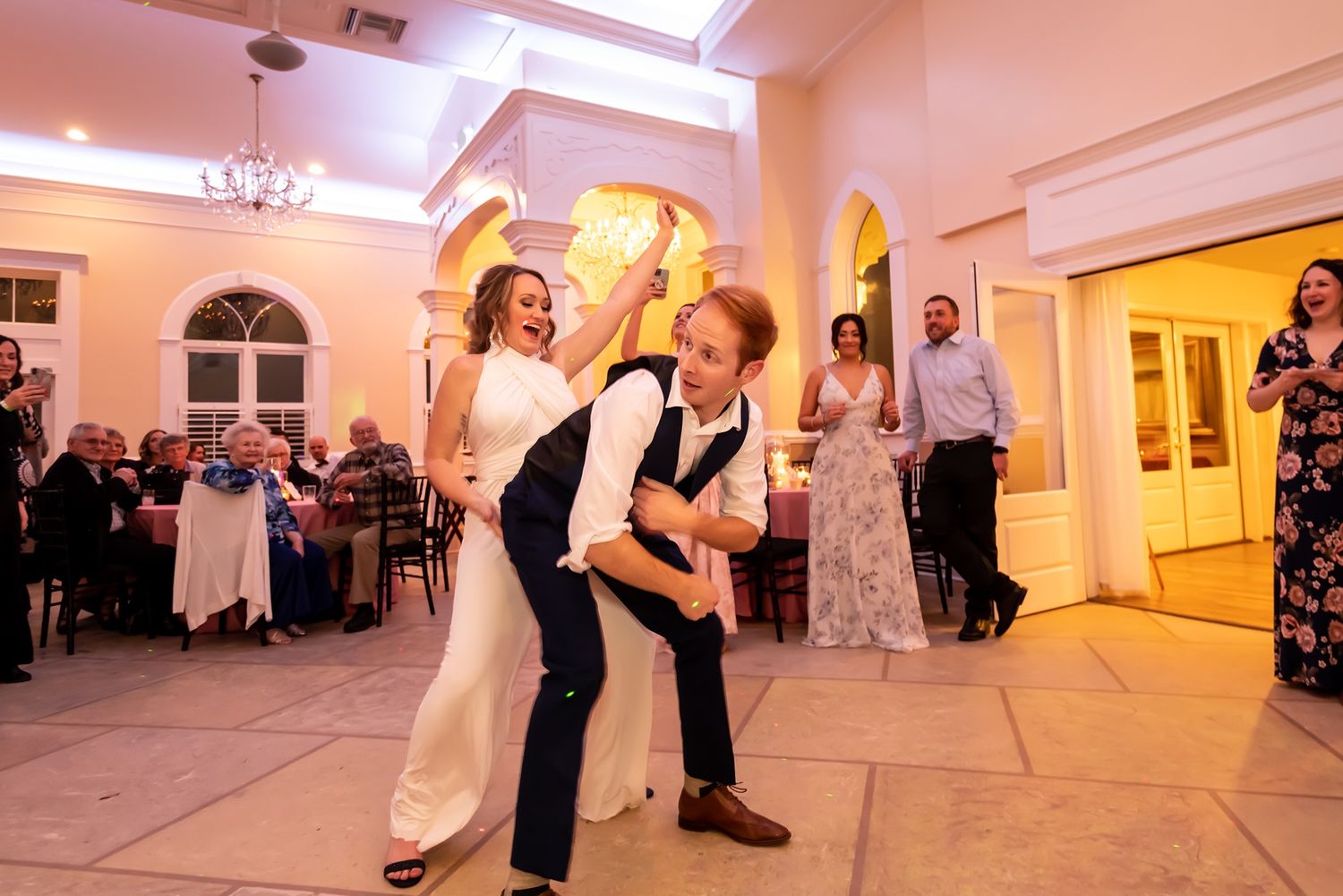 Wedding celebration with guests dancing in a spacious white reception hall with warm lighting.