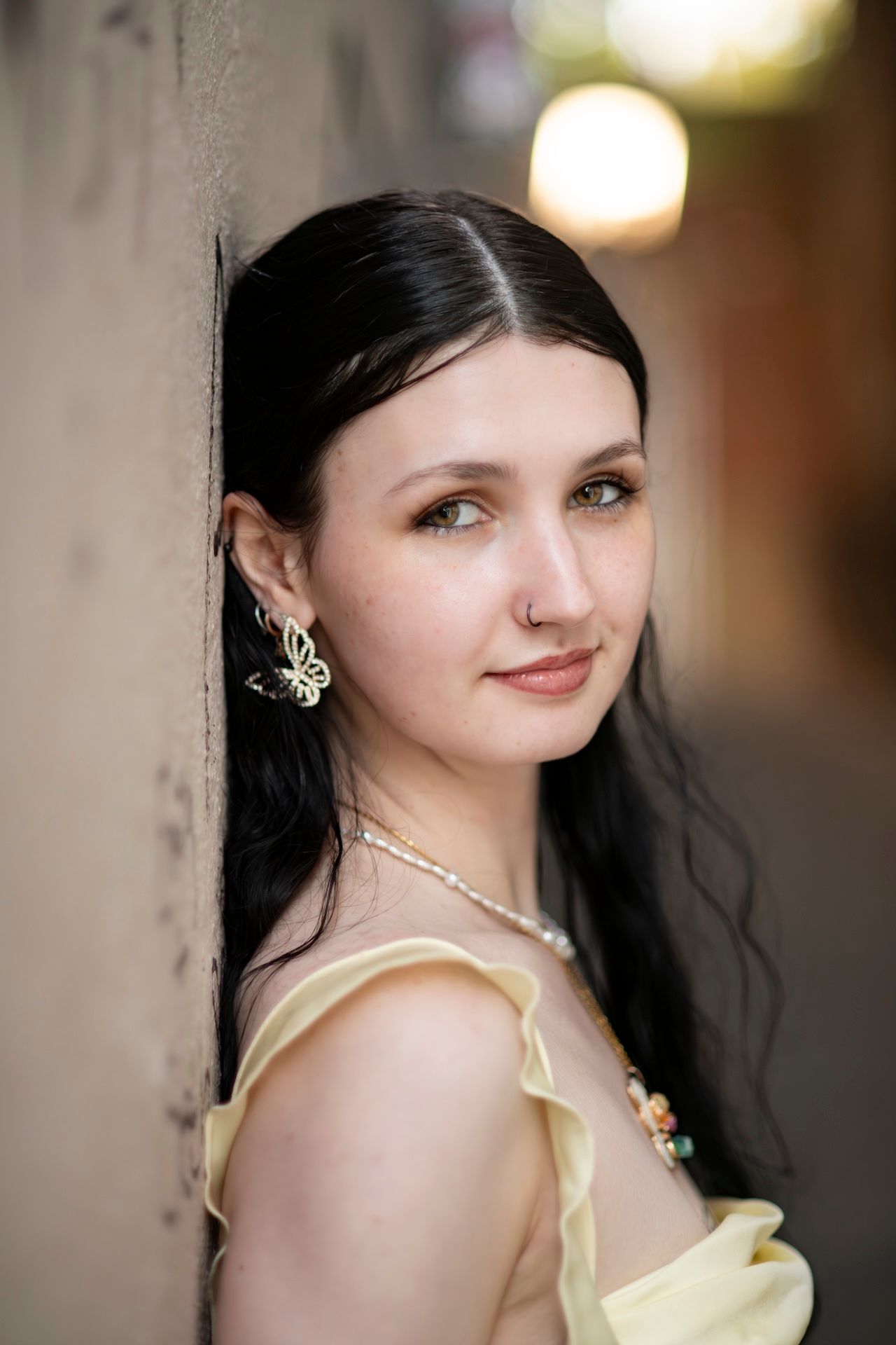 Young woman with dark hair in yellow dress, butterfly earrings, and necklace leaning against a wall outdoors.