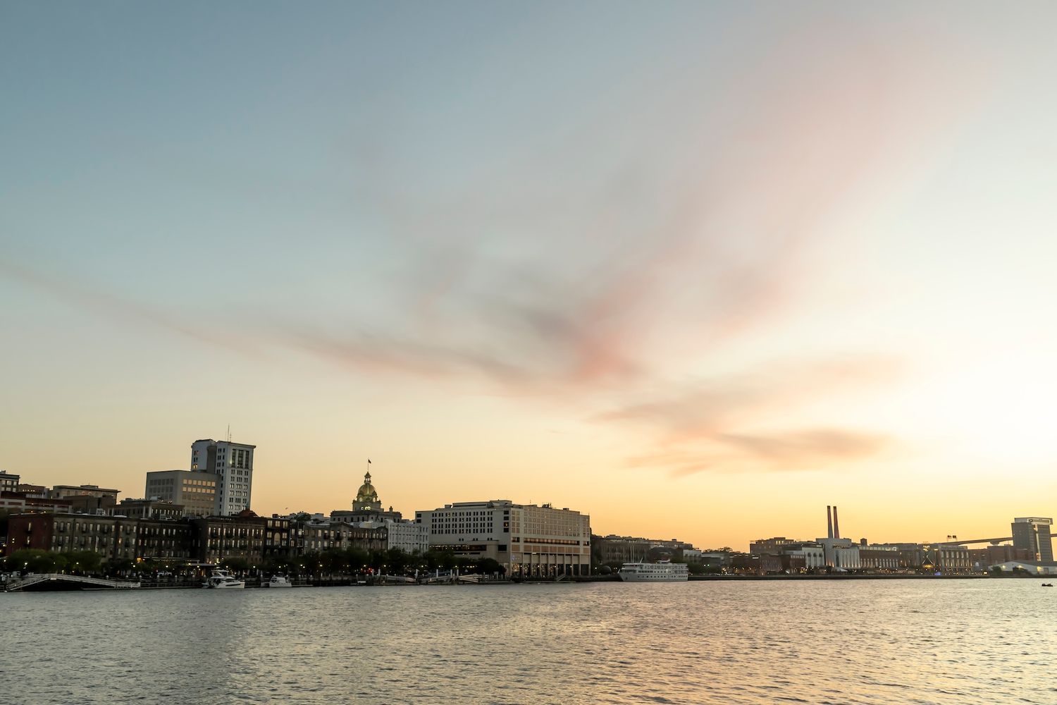 Panoramic waterfront cityscape at sunset with historic buildings and modern skyline reflecting warm golden hues over calm water.