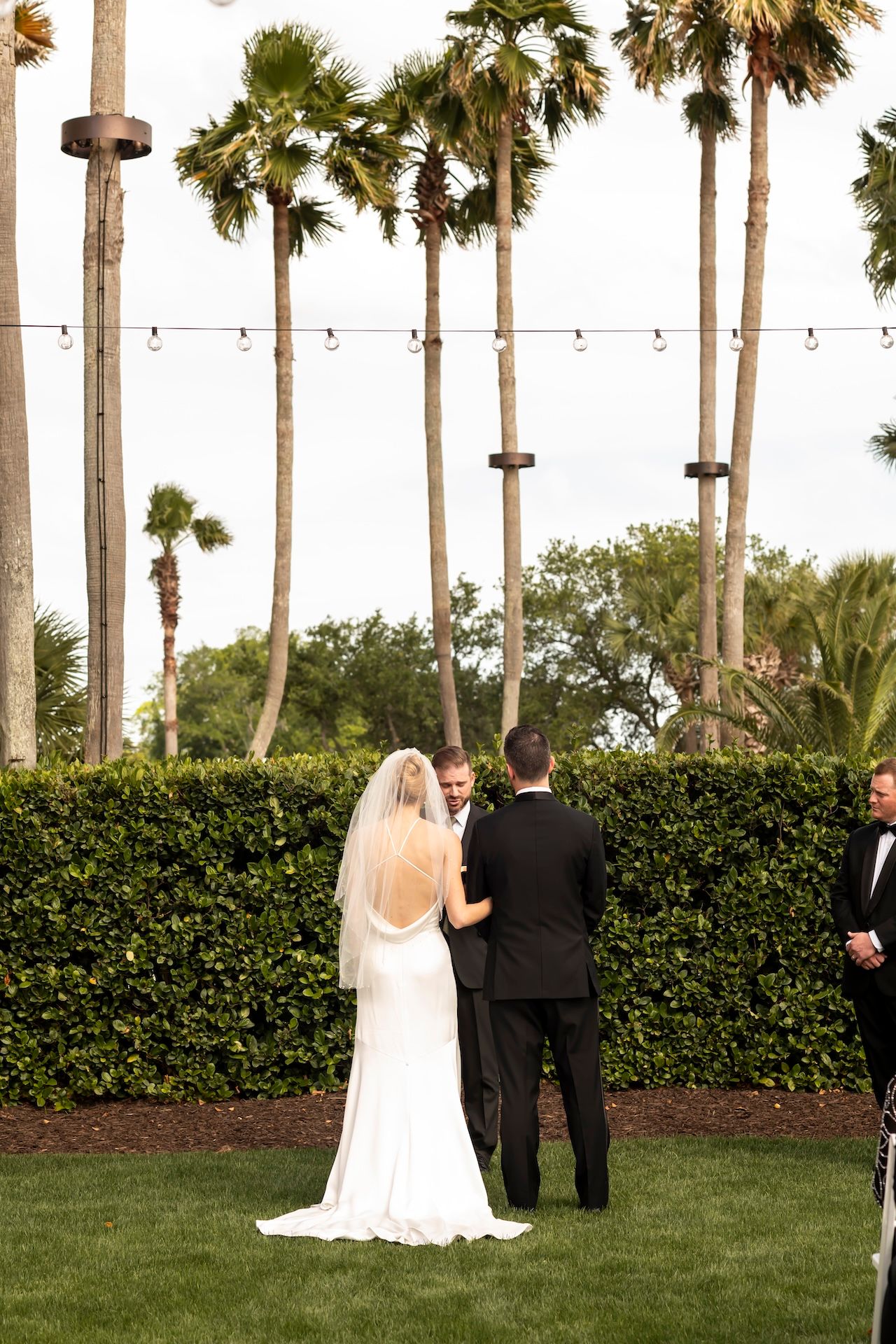 Bride and groom stand at outdoor wedding ceremony on green lawn surrounded by tall palm trees and string lights.