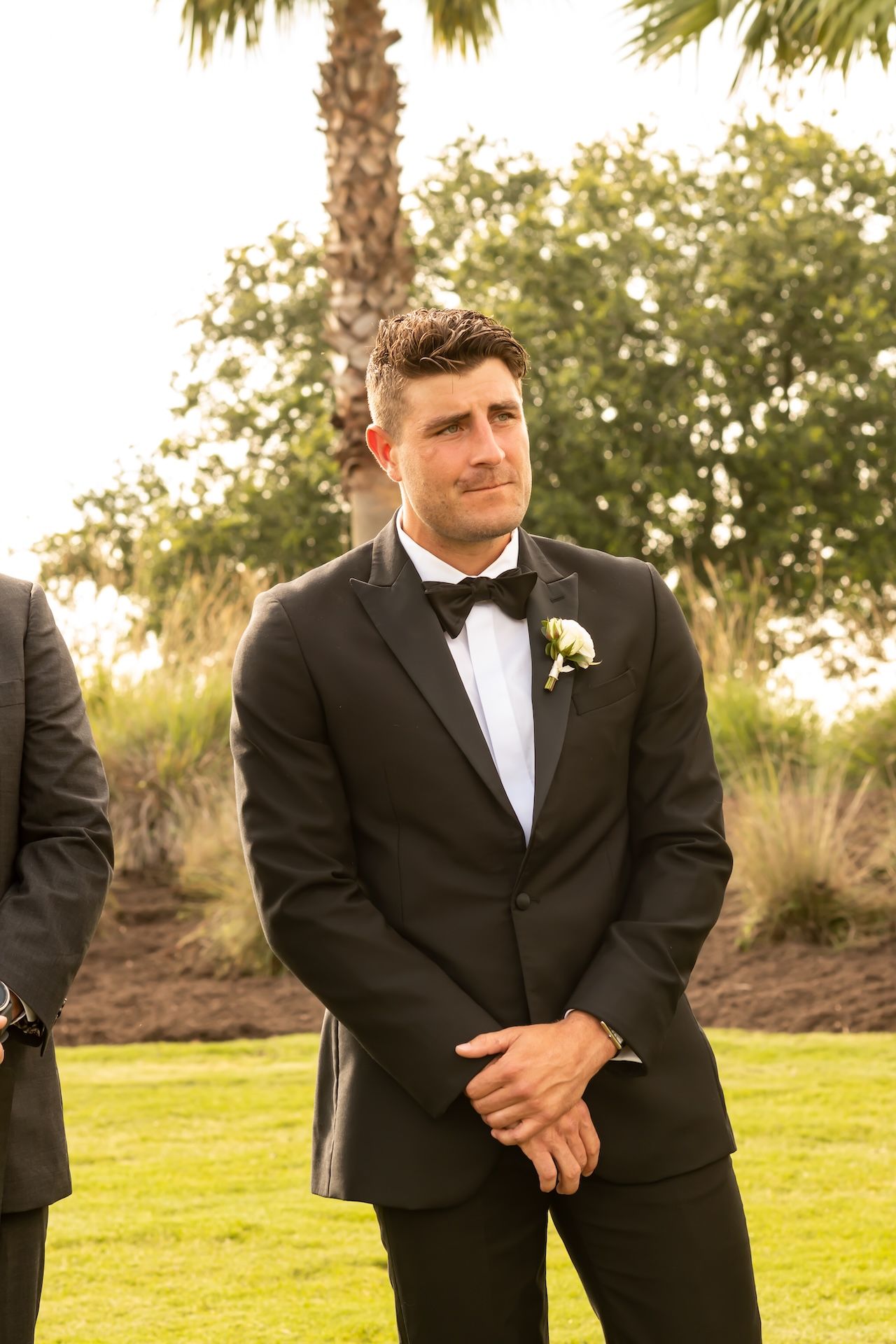 Groom in black tuxedo with bow tie and boutonniere stands outdoors, emotional during wedding ceremony near palm trees.