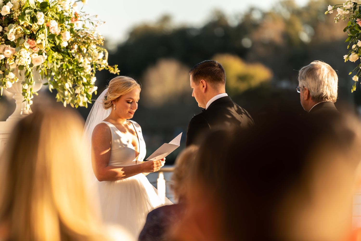 Bride reading wedding vows to groom at outdoor ceremony with floral arch and golden sunset light.