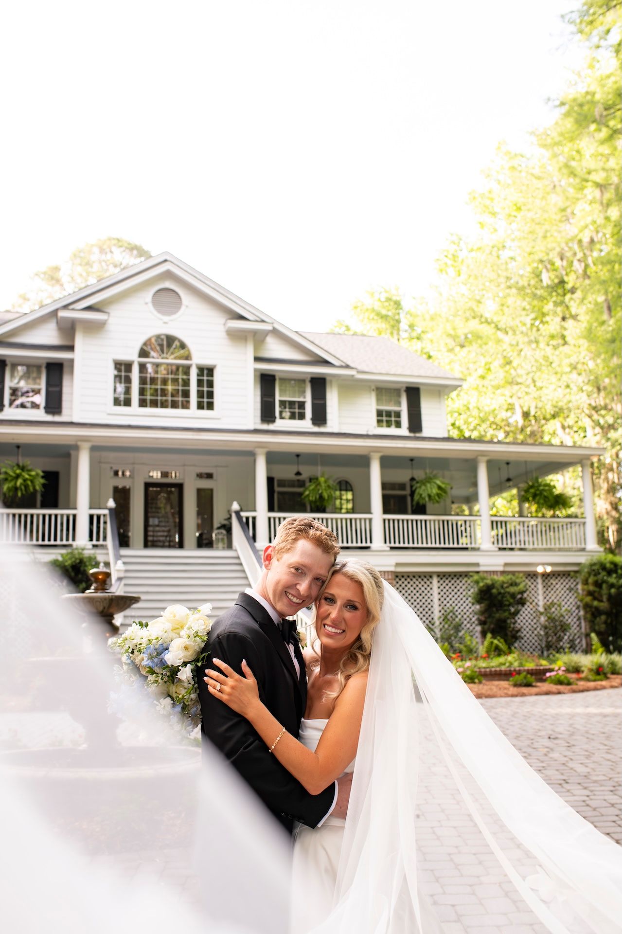 Bride and groom embrace in front of elegant white Southern mansion, bride's veil flowing, bouquet in hand.
