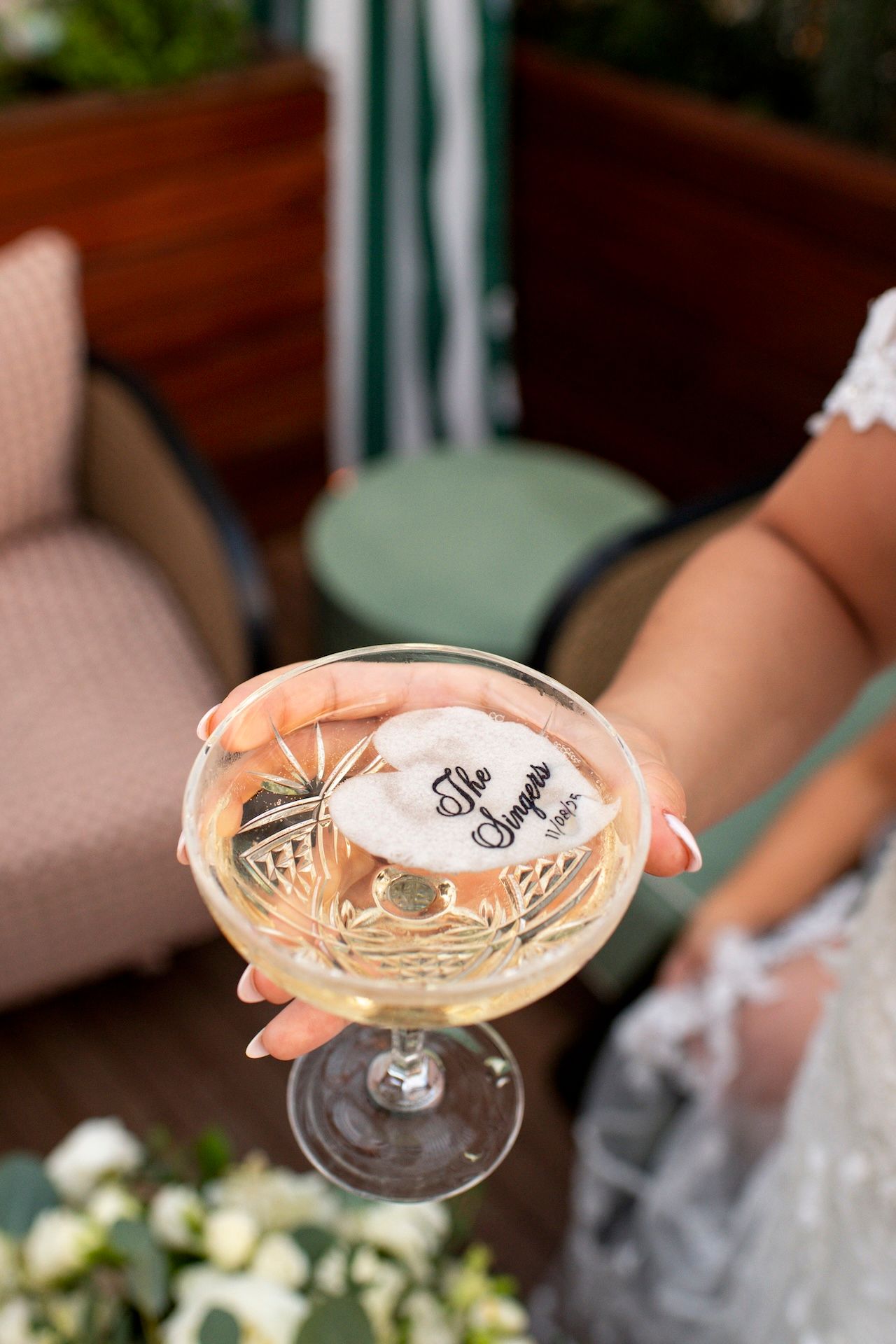 Several hands hold champagne coupes in a celebratory toast with green and floral decor in the background.