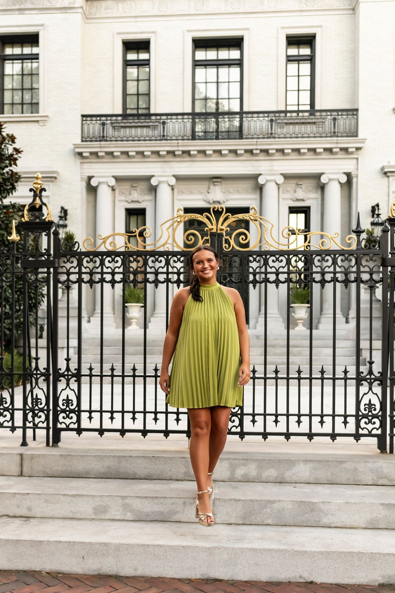 Woman in yellow sleeveless dress stands against backdrop of elegant white building with iron fencing.