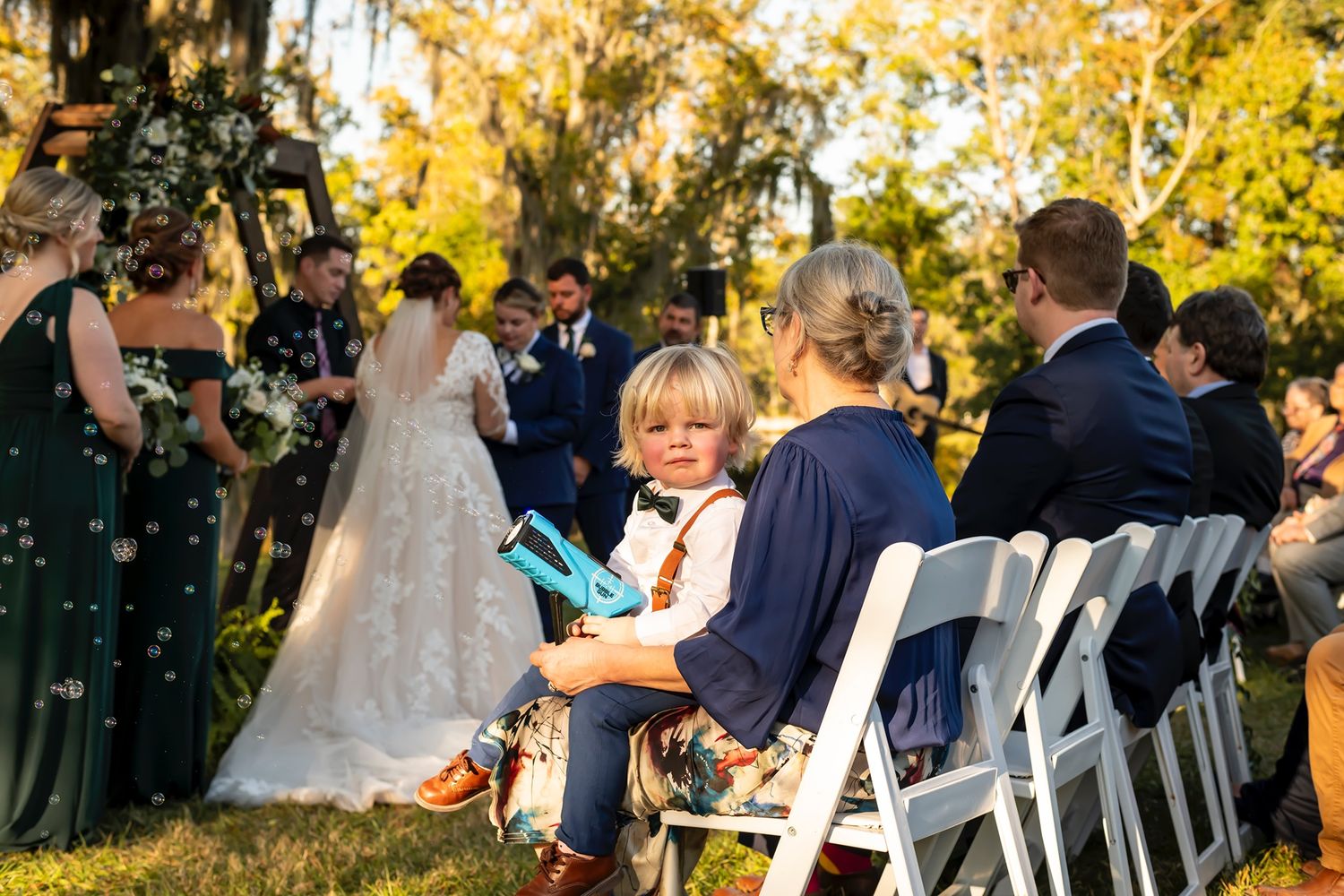 Young attendee plays handheld game during outdoor garden wedding ceremony with guests seated on white folding chairs.