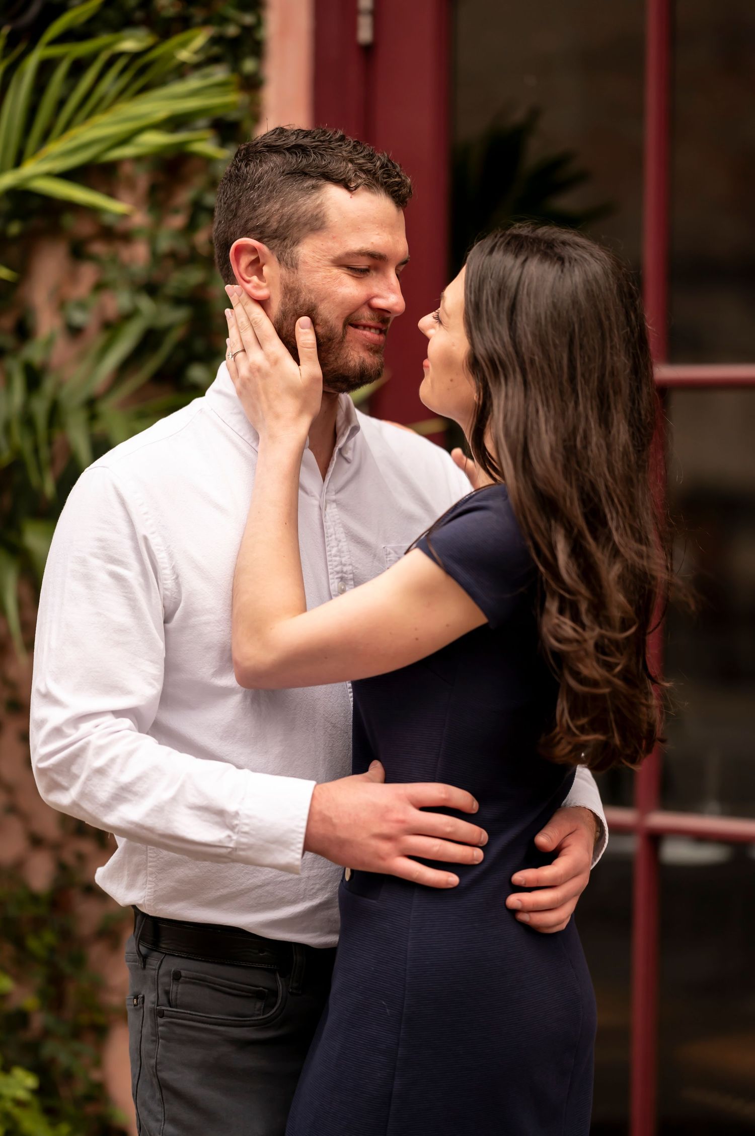 A romantic couple embraces outdoors, sharing an intimate moment in front of a red building with tropical foliage.