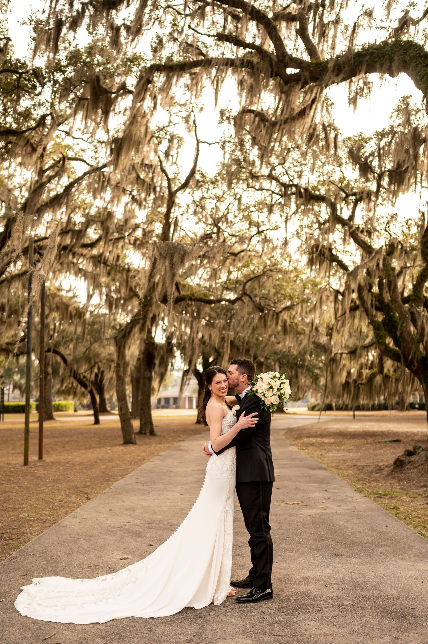 A couple embraces on a path beneath majestic Spanish moss-draped oak trees in a romantic outdoor setting.