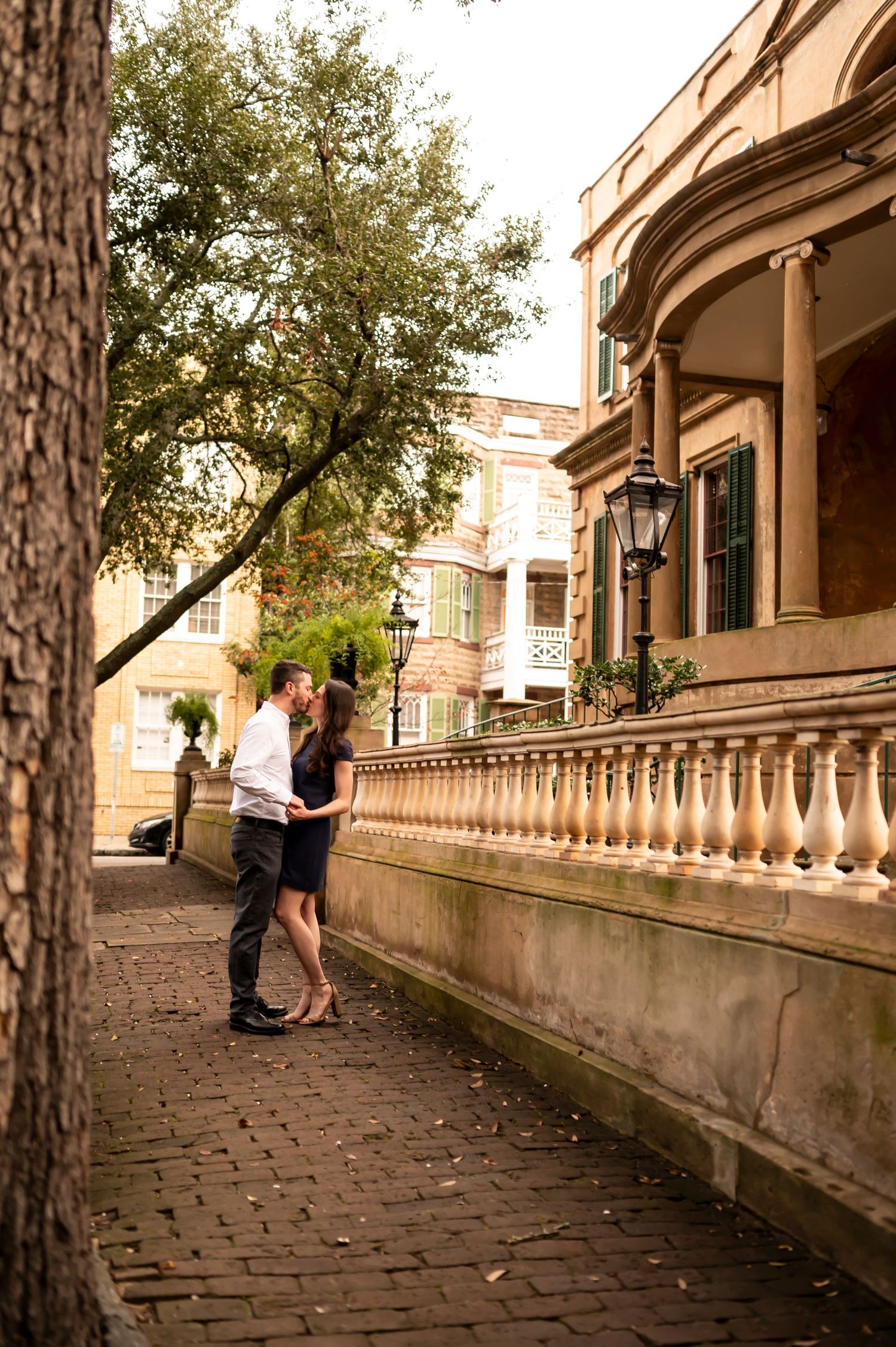 A couple shares an intimate moment on a historic brick walkway beside an elegant building with ornate balustrades and porches.