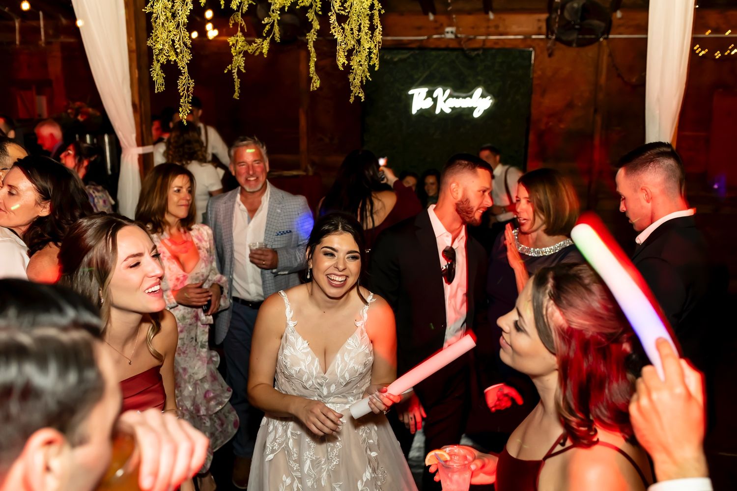 Wedding reception party with people dancing and celebrating under hanging greenery and neon lights in a rustic venue.