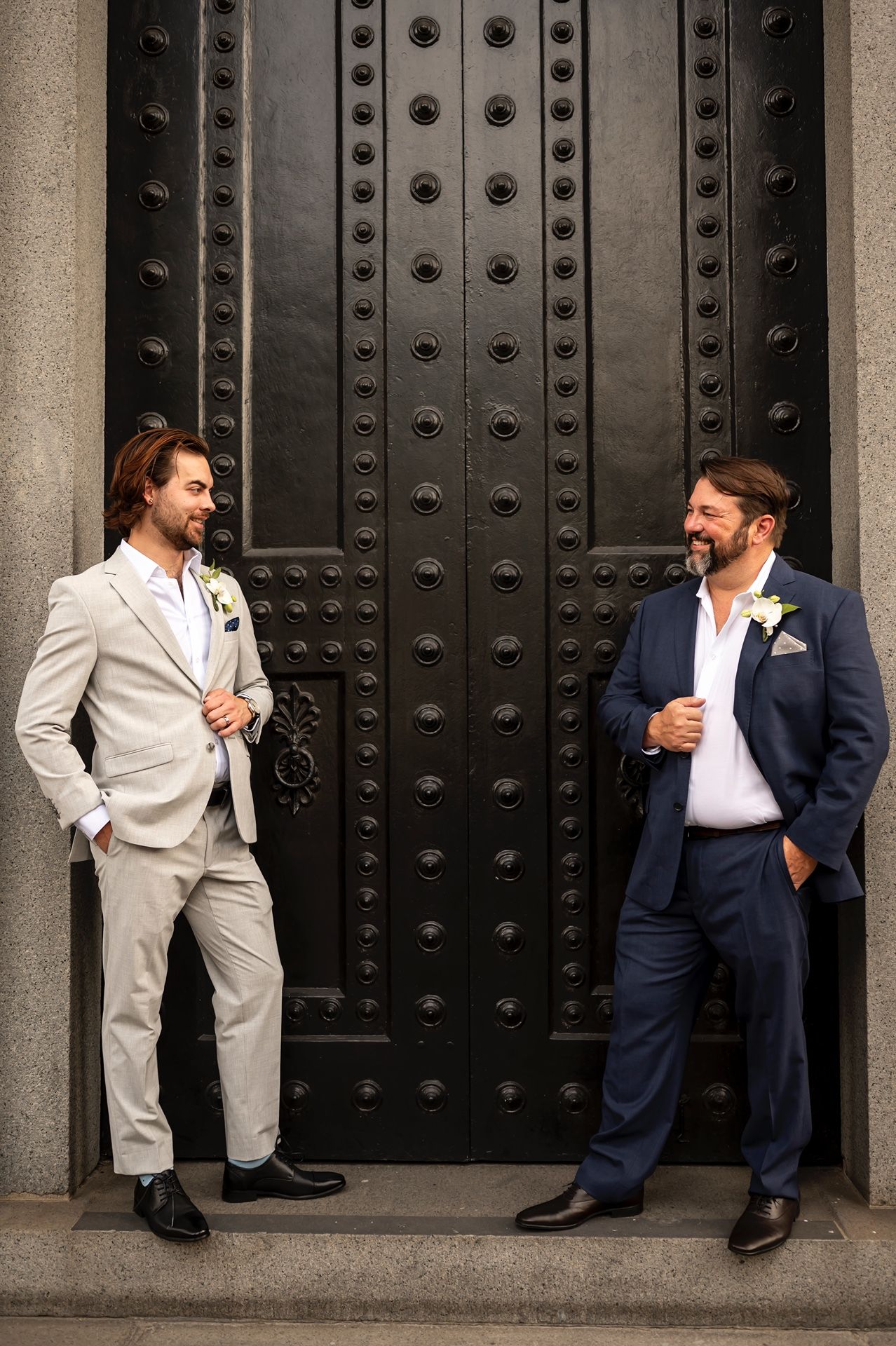 Two men in contrasting suits share playful interactions against a dark studded door backdrop.