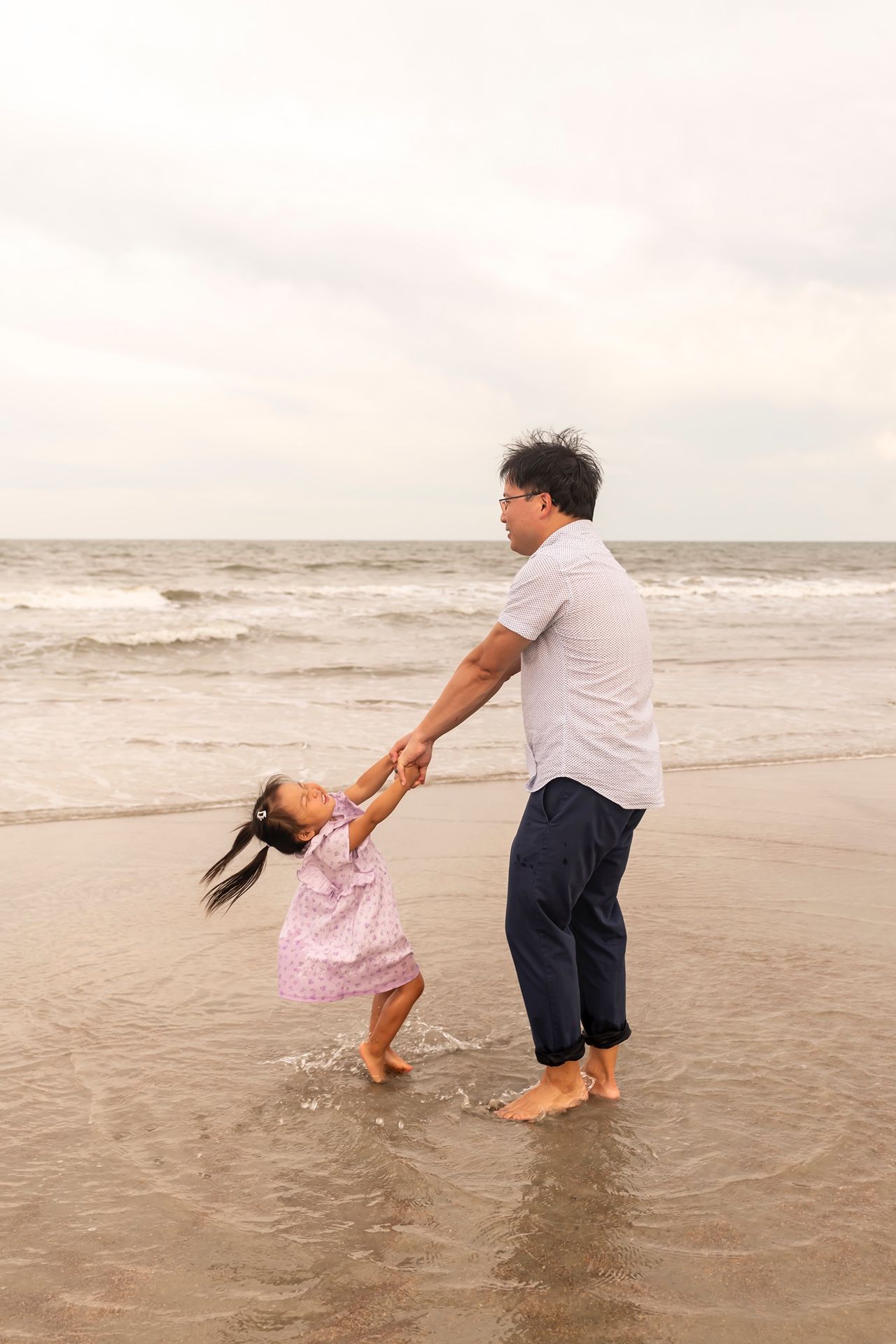 An adult and child in summer clothes play and dance together at the edge of the ocean on a sandy beach.