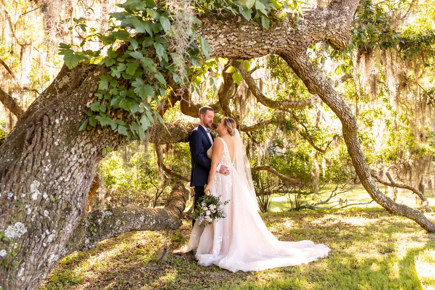 A romantic wedding couple stands hand in hand beneath sun-dappled oak trees draped in Spanish moss.