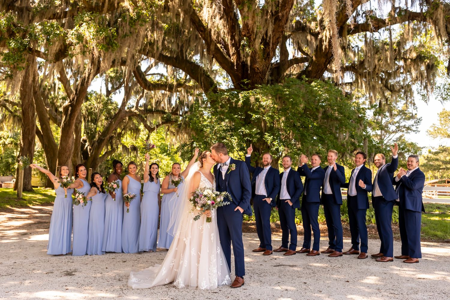 Wedding party in navy suits and light blue dresses poses together under Spanish moss draped trees for outdoor photos.