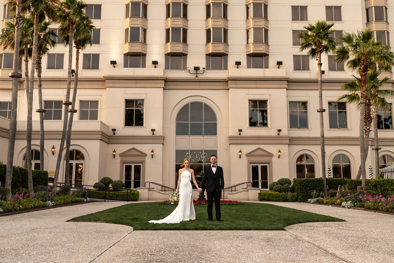 A romantic wedding photo in front of an elegant beige hotel building with palm trees and manicured gardens.