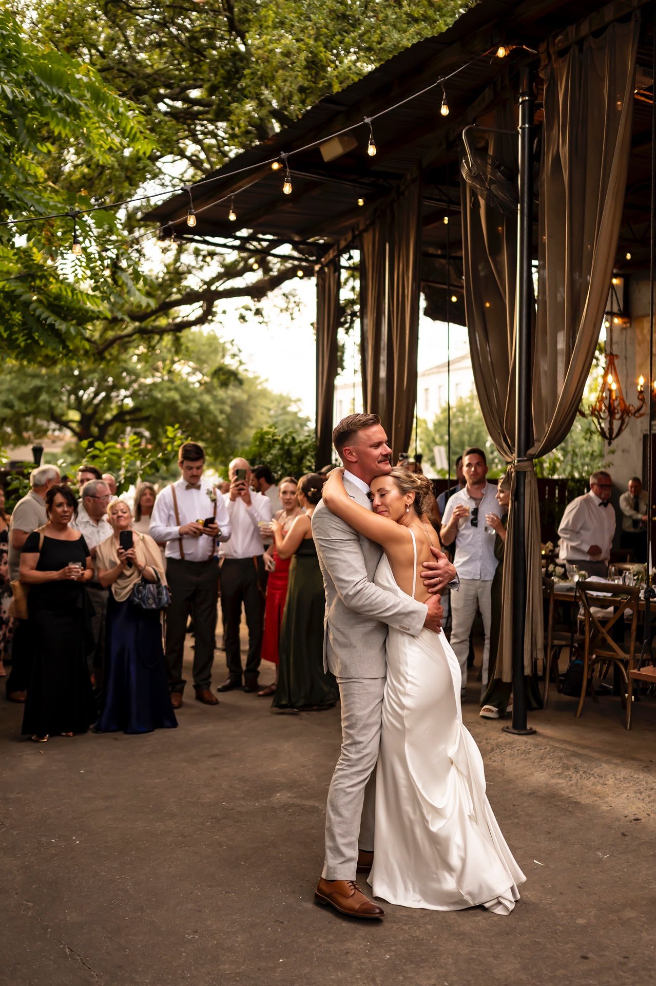 A couple shares their first dance under string lights at a rustic outdoor wedding venue while guests watch in celebration.