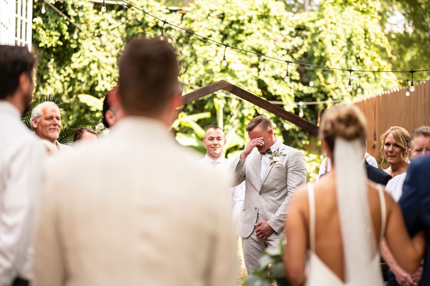 Wedding guests toss flower petals at a happy couple during an outdoor celebration in a sunlit garden.