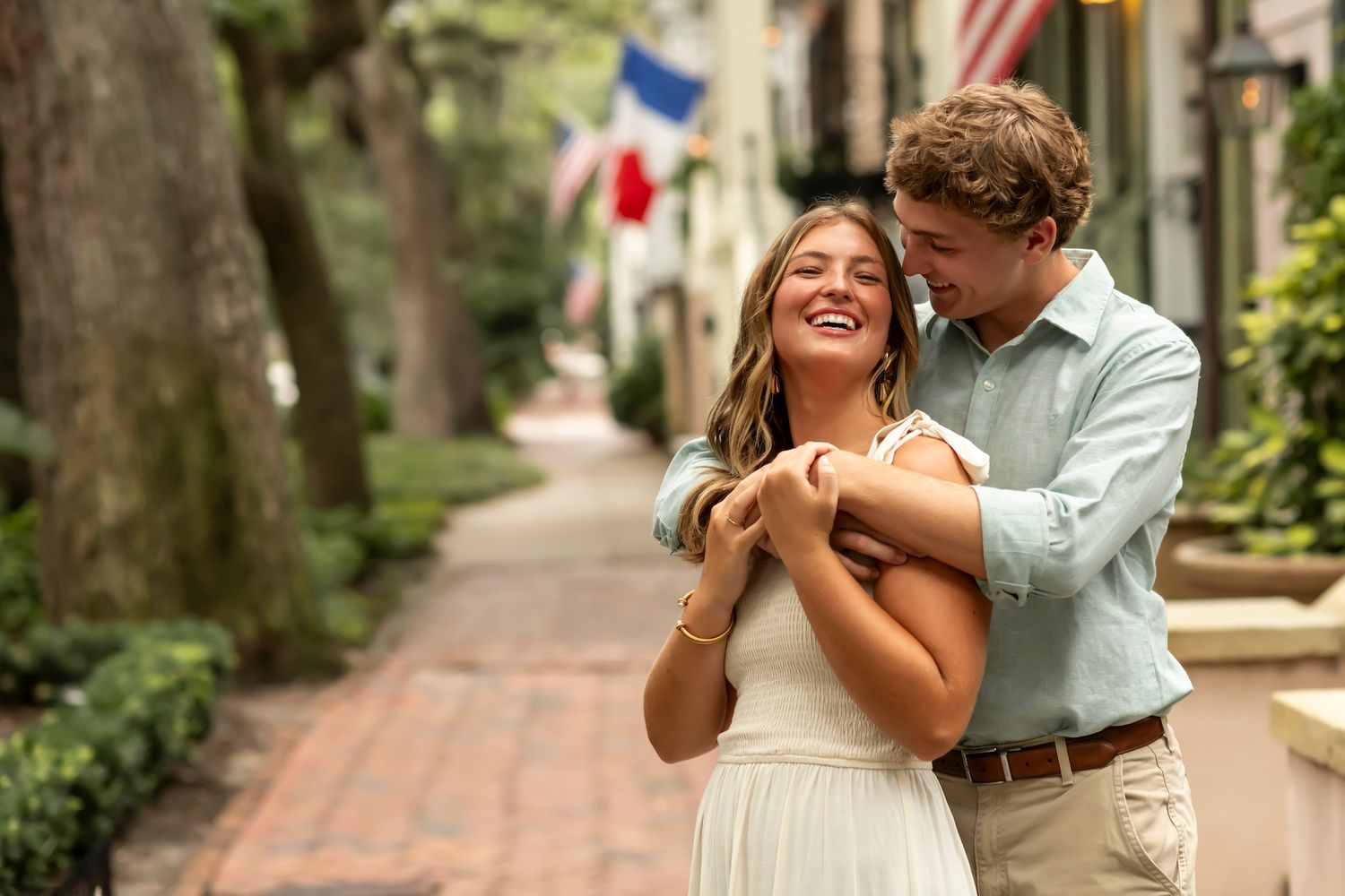 A couple in casual wedding attire embrace and share a tender moment on a brick-lined street with flags overhead.