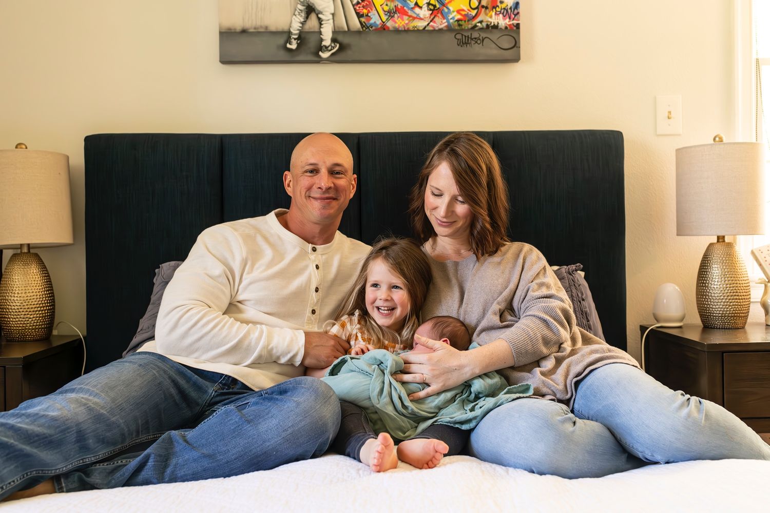 Family relaxes together on a grey couch with decorative lamps and artwork visible in background.