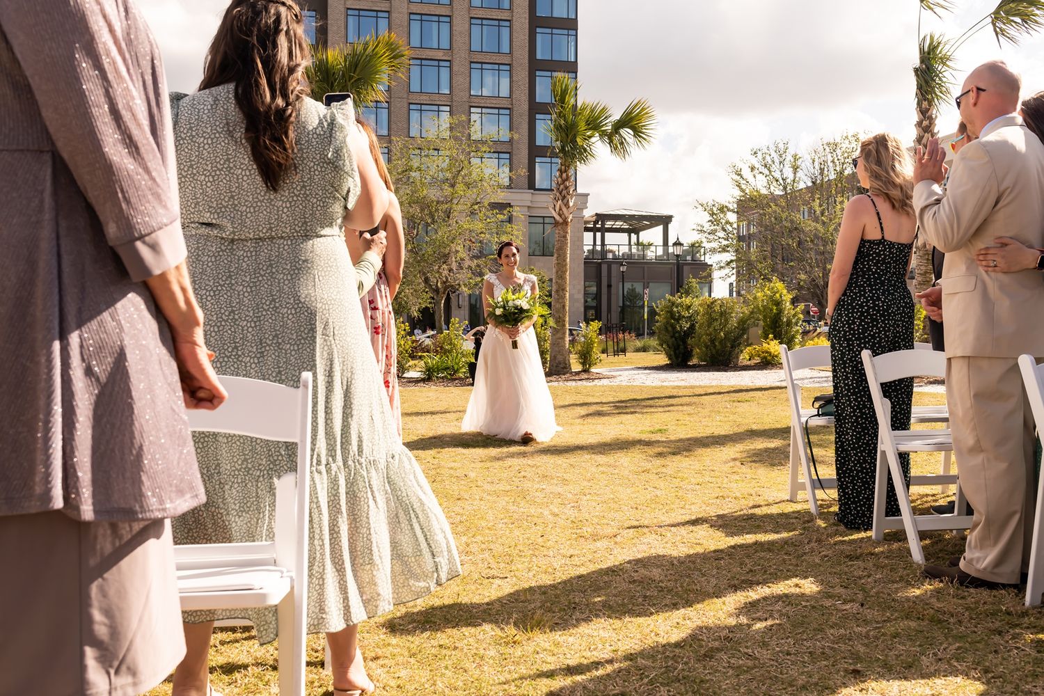 Wedding guests in light-colored attire mingle outdoors on a sunny day near modern apartment buildings.