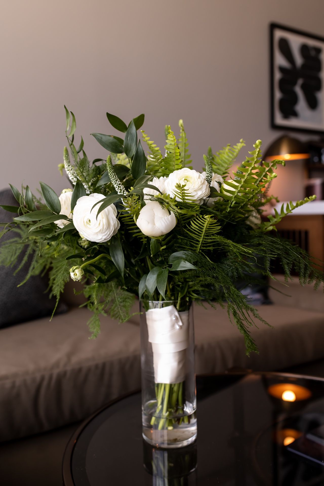 A white peony and fern bouquet arranged in a clear glass vase sits atop a dark coffee table with candlelight ambiance.